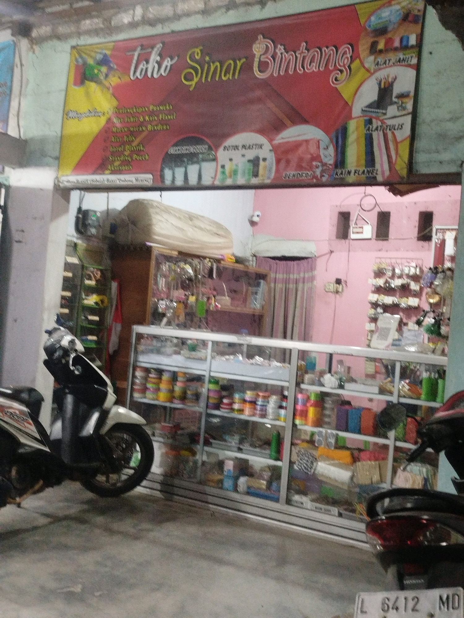 The image shows the storefront of "toko Sinar Bintang" in Burneh, Indonesia. The store appears to be a retail establishment selling various craft and art supplies, as indicated by the banner displaying "ALAT JAHIT" (sewing tools), "ALAT TULIS" (writing tools), and "KAIN FLANEL" (flannel fabric). In front of the store, two motorcycles are parked. The store's interior is visible through glass display cases filled with colorful spools of thread, fabric, and other craft materials. The exterior of the building has a pink painted wall. The overall scene depicts a typical small business in an Indonesian town.