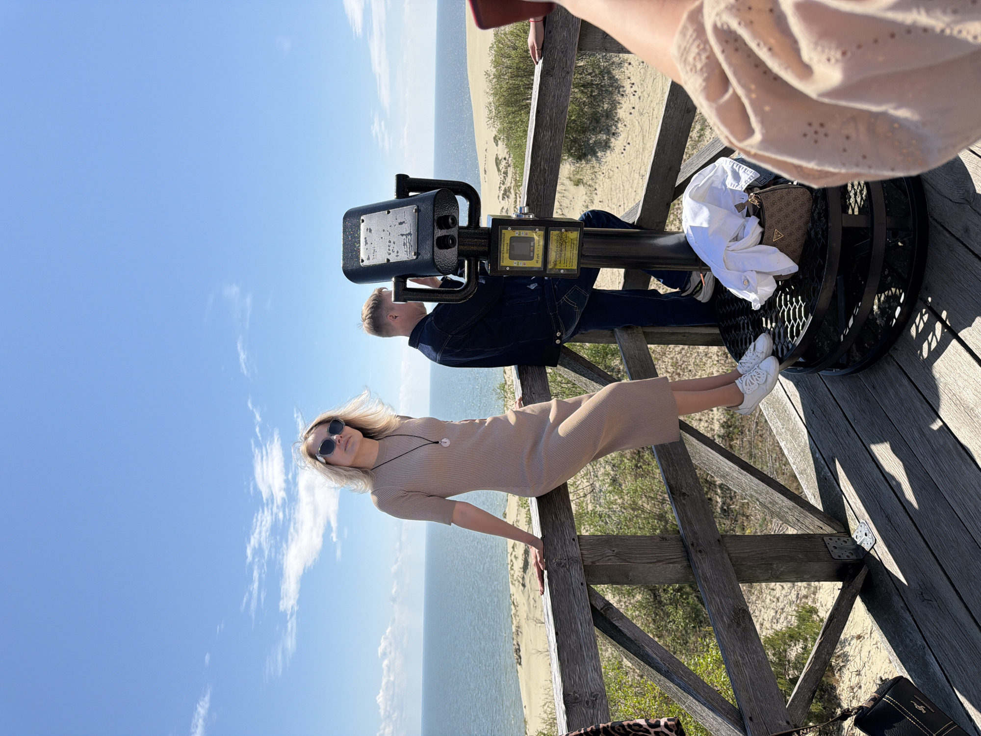The image captures a scene on a wooden observation deck overlooking a sandy landscape and the sea under a clear blue sky with scattered clouds. In the foreground, a woman with blonde hair and sunglasses is leaning on a wooden railing, wearing a beige knit dress and white sneakers. To her right, a man in a denim jacket and jeans is looking through a large telescope. A small, patterned handbag and a white shirt are placed on the base of the telescope. In the far right, a portion of another person's arm and shoulder, wearing a light-colored, textured garment, is visible. The environment suggests a coastal or dune area, possibly within the vicinity of Morskoye, Russia, known for its sand dunes.
