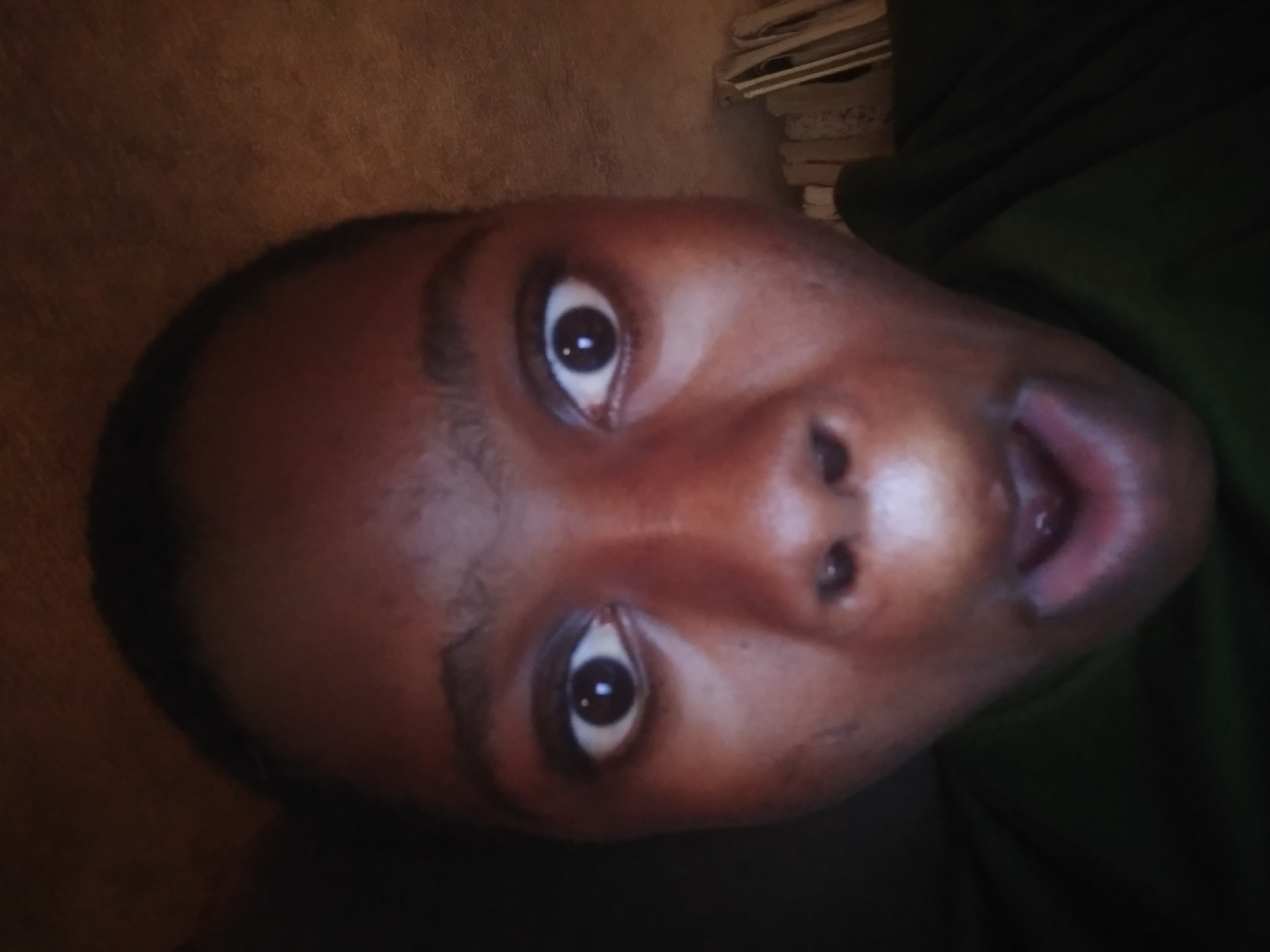 A close-up, rotated perspective photograph depicts an individual's face. The subject has brown skin, dark eyes, and an open mouth. They appear to be wearing a dark green garment. The background shows a beige surface and a stack of papers or books. The location is unknown.