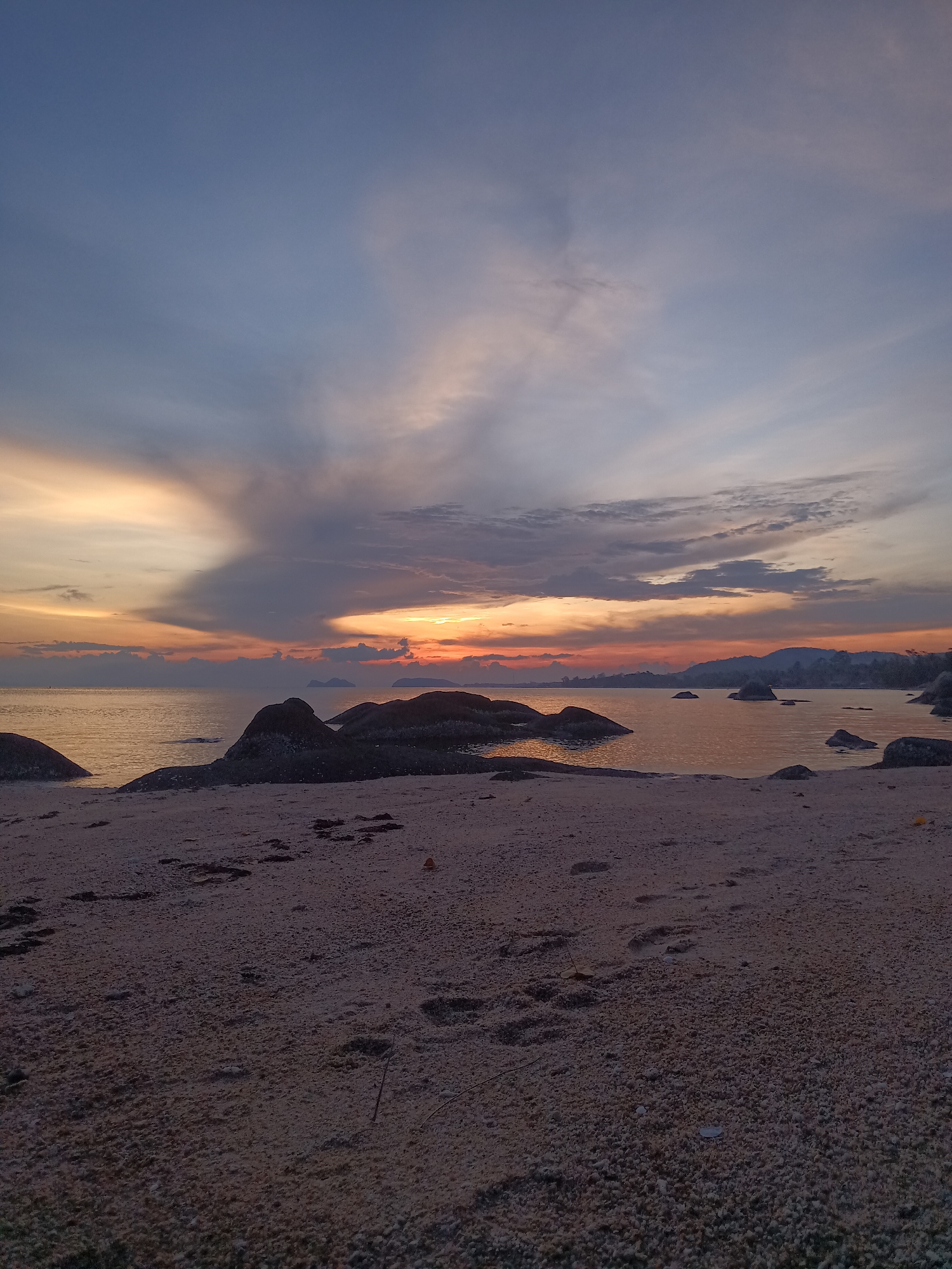 A wide shot captures a serene scene at Baan Kai, Thailand. The foreground is dominated by a sandy beach, exhibiting numerous footprints. The middle ground features a calm body of water, dotted with several rock formations. The horizon reveals a coastline silhouette. Above, a dramatic sky showcases a blend of blue, gray, and orange hues, indicative of either sunrise or sunset.