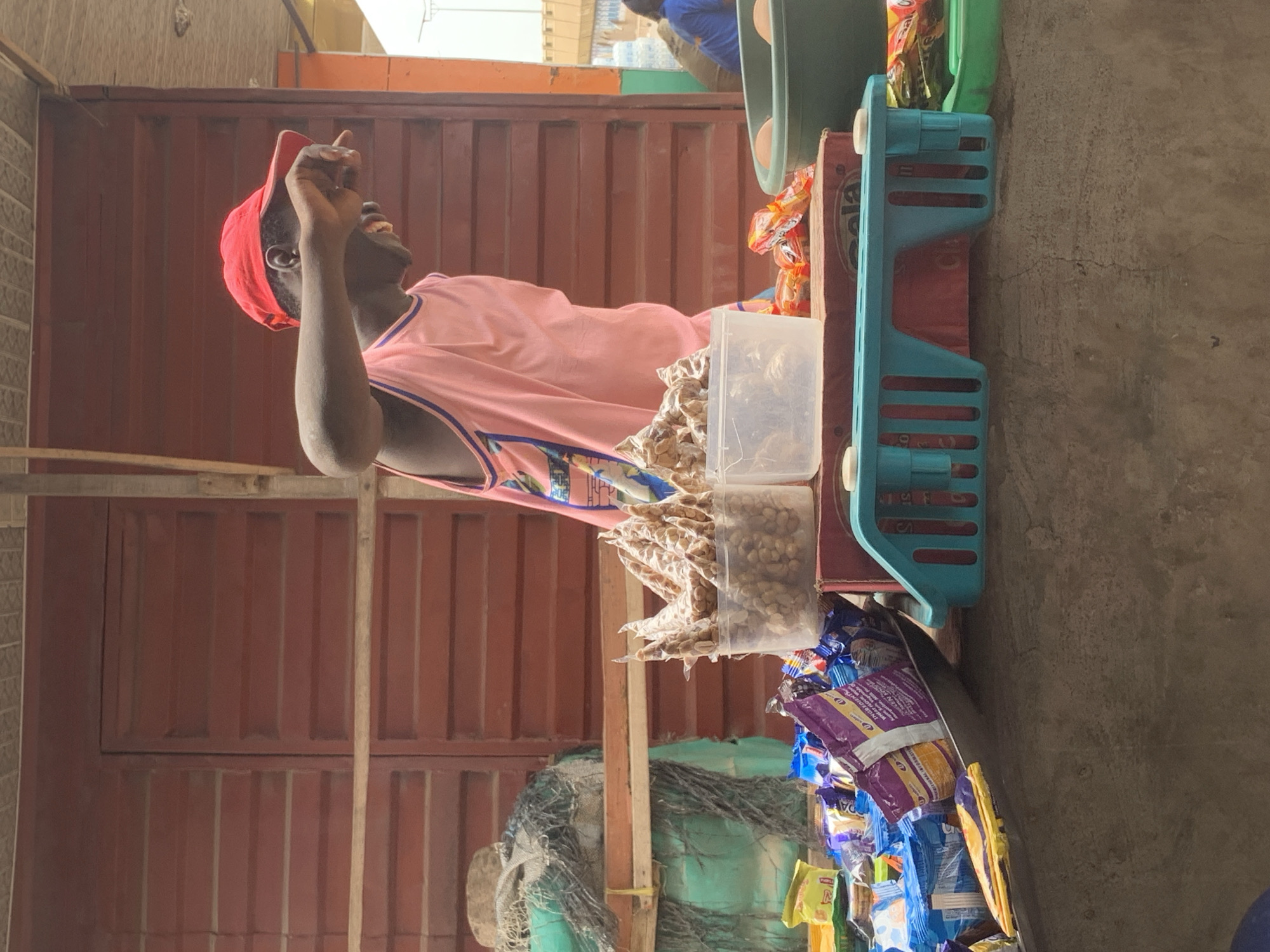 A young Black male wearing a red baseball cap and a pink tank top with blue trim stands at a market stall in Shagamu, Nigeria. He is partially obscured from the chest up, with his right arm extended and his hand touching his face as if shielding his eyes from the sun or waving. The stall is laden with various goods. In the foreground, an array of packaged snacks and candies, mostly in blue, purple, and yellow wrappers, are piled on a metal tray and a concrete surface. Behind these, a clear plastic container holds small, individually wrapped portions of what appear to be nuts or fried snacks. To the right, a green basin holds two eggs, and next to it are more colorful snack packages. A red crate forms a base for some of these items. The stall's backdrop is a corrugated red metal wall with horizontal slatted sections. To the left, a large, green-wrapped object, possibly a bundle of goods or a storage container, is covered with netting and twine. The overall scene depicts a vibrant market environment with a focus on food items.