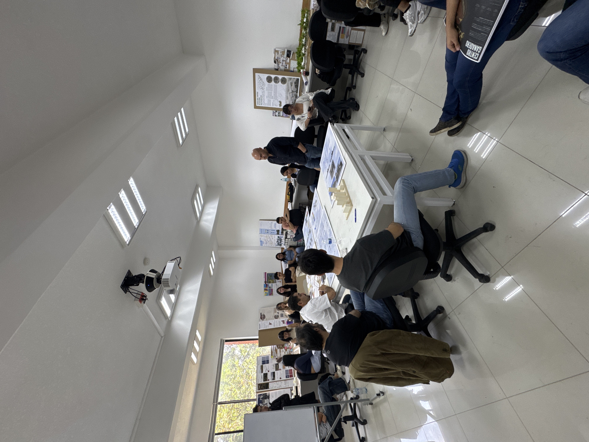 The image shows an indoor scene, apparently a classroom or studio, in San Andrés Cholula, Mexico. There are approximately 20-25 people present, most of whom appear to be students. They are gathered around several white tables on which architectural models and drawings are displayed. An older man, possibly an instructor, stands at one of the tables and appears to be lecturing or giving feedback. Most of the students are seated in chairs, listening or observing. Some are leaning forward, engaged with the presentation. In the background, there are posters and architectural drawings displayed on the walls. The room has white walls, tiled floors, and large windows that let in natural light. A projector is mounted on the ceiling. The atmosphere seems to be one of a design review or critique session.