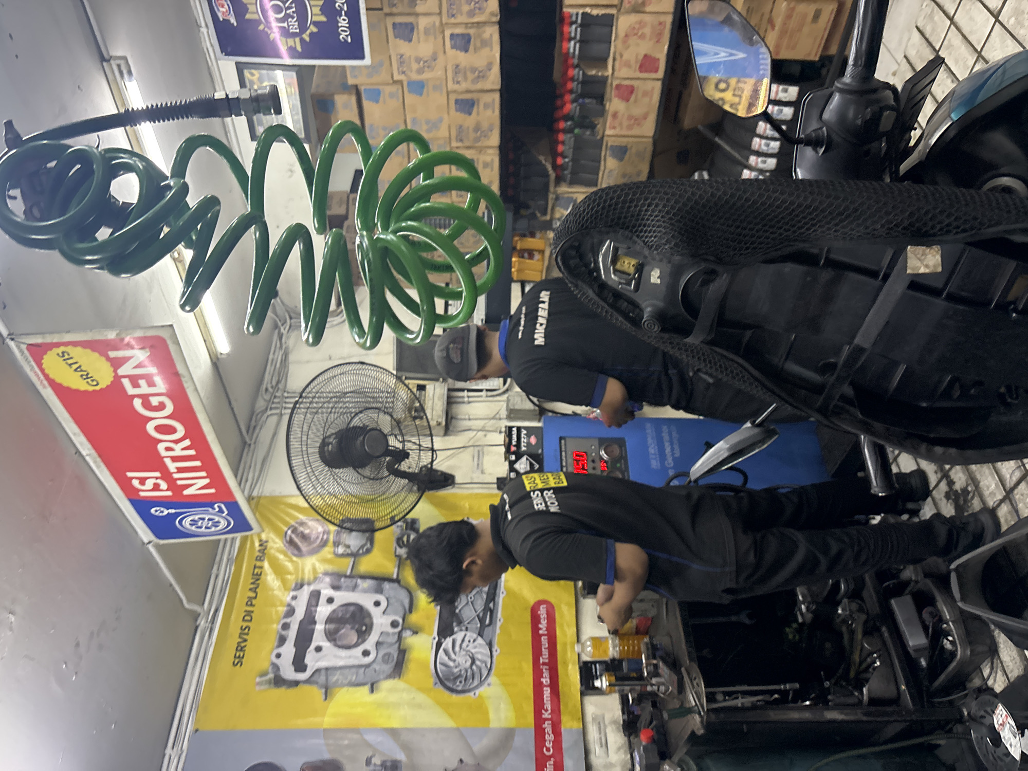 The image depicts the interior of a motorcycle service shop, likely located in North Jakarta, Indonesia, given the signage. Two men, dressed in black shirts and pants, are engaged in servicing a motorcycle. One man is standing behind the motorcycle, facing away from the camera, working on a component. The other man is facing the camera, also working on the motorcycle. Various tools and equipment are visible in the foreground and background, including a workbench, a fan, and shelves stocked with parts and fluids. A prominent banner in the background advertises "Isi Nitrogen" (Fill Nitrogen) and "Servis di Planet Ban" (Service at Planet Ban), which is a known tire service chain in Indonesia. A coiled green air hose hangs from the ceiling. The environment appears to be a well-lit, functional workshop.