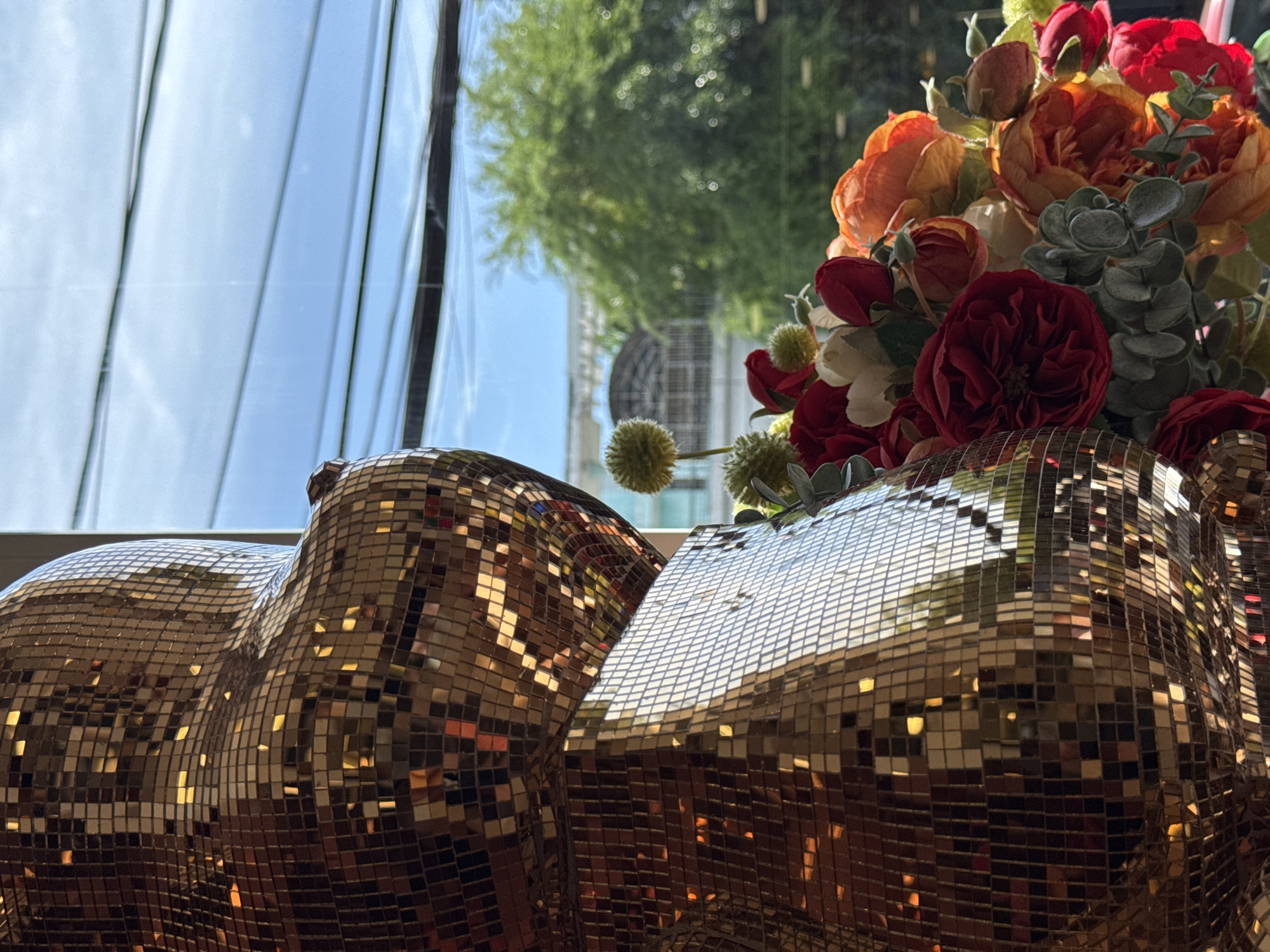 A close-up shot captures the reflective surface of a large, gold, mosaic-covered bear statue in the foreground. To the right of the statue, a vibrant bouquet of artificial flowers, including red roses, orange peonies, and greenery, is arranged. In the background, visible through a window, is a bright blue sky with some power lines strung across it. Beyond the power lines, trees with lush green foliage and a building with an arched structure are discernible. The scene appears to be set indoors, with the window offering a view of an outdoor environment in Nha Trang, Vietnam. There are no people or animals present in the image.