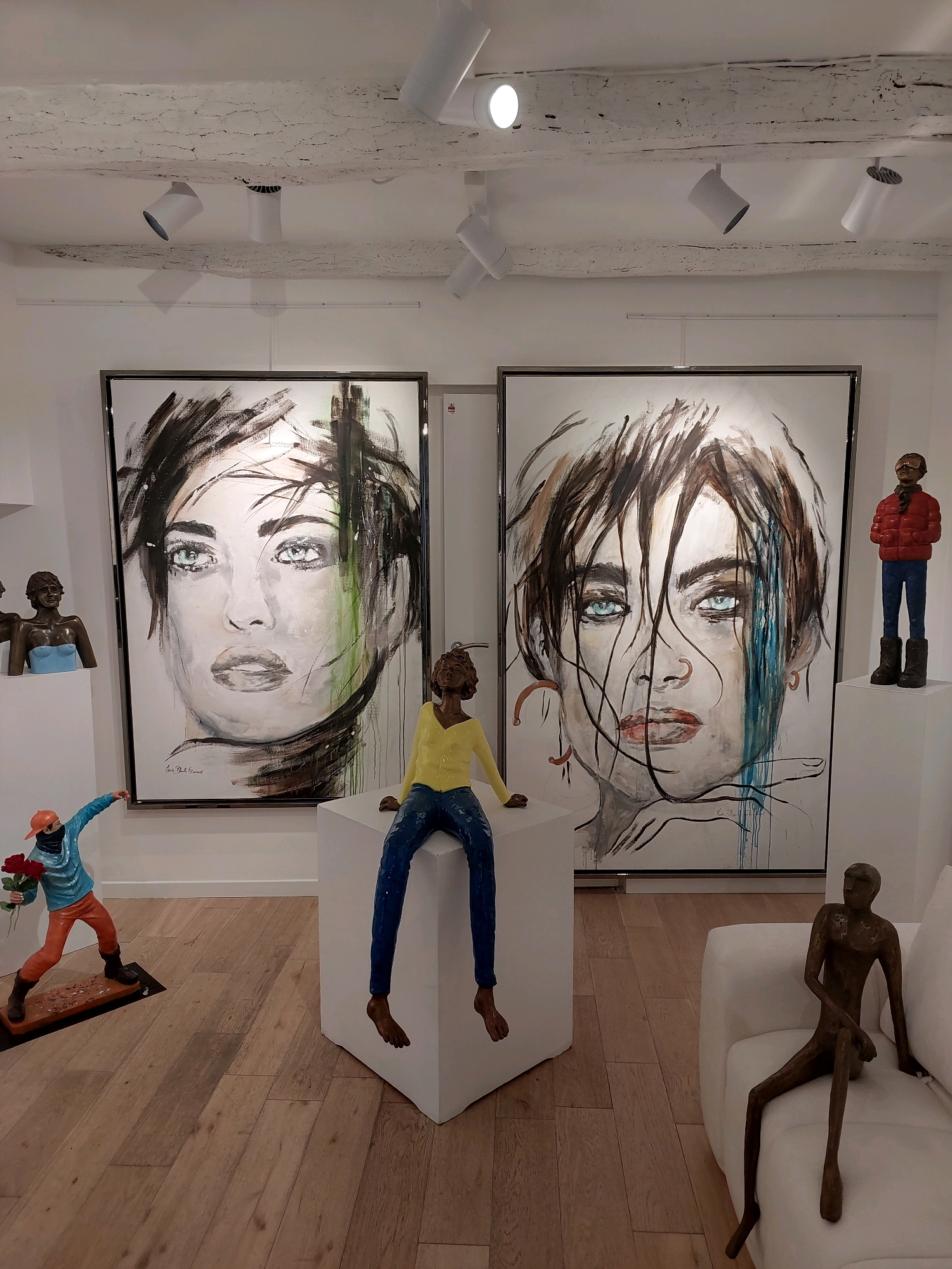 The image depicts the interior of an art gallery in Saint-Paul-de-Vence, France. Two large framed paintings of women's faces with striking blue eyes are prominently displayed on a white wall. The painting on the left features a softer, more diffused rendition of the face, while the one on the right is more detailed and intense, with visible brushstrokes and dripping paint.
In the foreground, several sculptures add a three-dimensional element to the scene. On the left, a sculpture of a person in a blue jacket and orange pants, holding a bouquet of red roses, stands on a dark base. To the right of the paintings, a white cube pedestal supports a sculpture of a seated figure wearing a yellow top and blue pants. Further to the right, a bronze sculpture of a slender, seated figure rests on a white sofa.
The gallery space features exposed wooden beams on the ceiling, from which multiple track lights are suspended, illuminating the artwork. The floor is made of light-colored wooden planks. The overall ambiance suggests a contemporary art exhibition space.