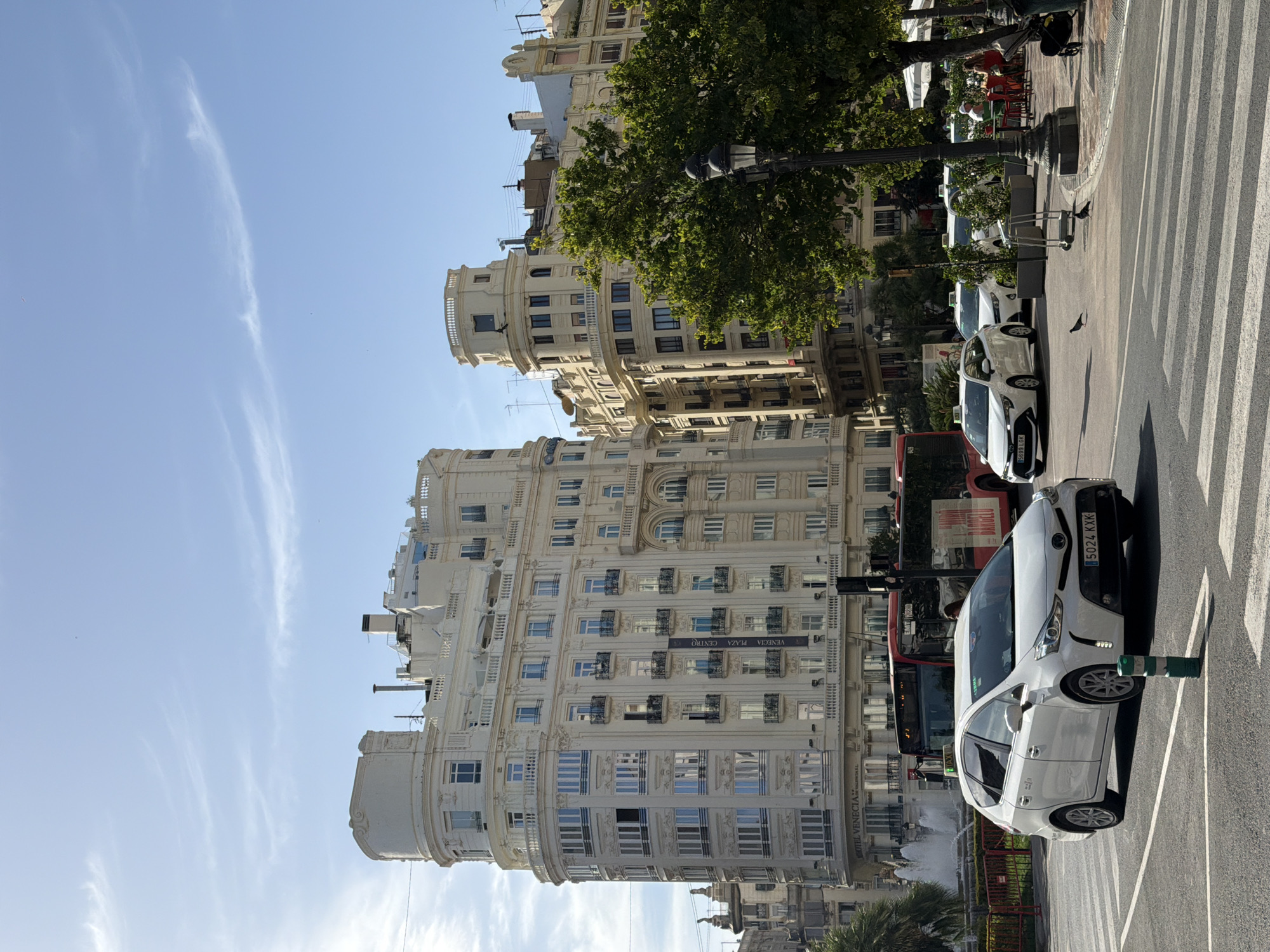 The image captures a street scene in the city of València, Spain, likely in the vicinity of Plaça de l'Ajuntament, given the architectural style and presence of a bus stop with signage that could be associated with public transport hubs in the city. In the foreground, a white Toyota Prius taxi, license plate 5024 XKX, is turning at an intersection with a pedestrian crossing. Another white taxi is partially visible to the right of the Prius. Behind the taxis, a red bus is stopped, possibly at a bus stop, with people visible inside.
Dominating the background are several large, ornate white buildings with multiple stories, featuring numerous windows, balconies with wrought-iron railings, and decorative architectural elements. One building prominently displays signage that reads "HOTEL VENECIA" and "VALENCIA PLAZA CENTRO," confirming the location. A fountain is visible to the far left, with water jets spraying upwards. Green trees line the right side of the street, with tables and chairs set up at an outdoor cafe area beneath them, where a few people are seated. The sky is blue with scattered light clouds, suggesting a sunny day. The overall impression is of a lively urban square in a European city.