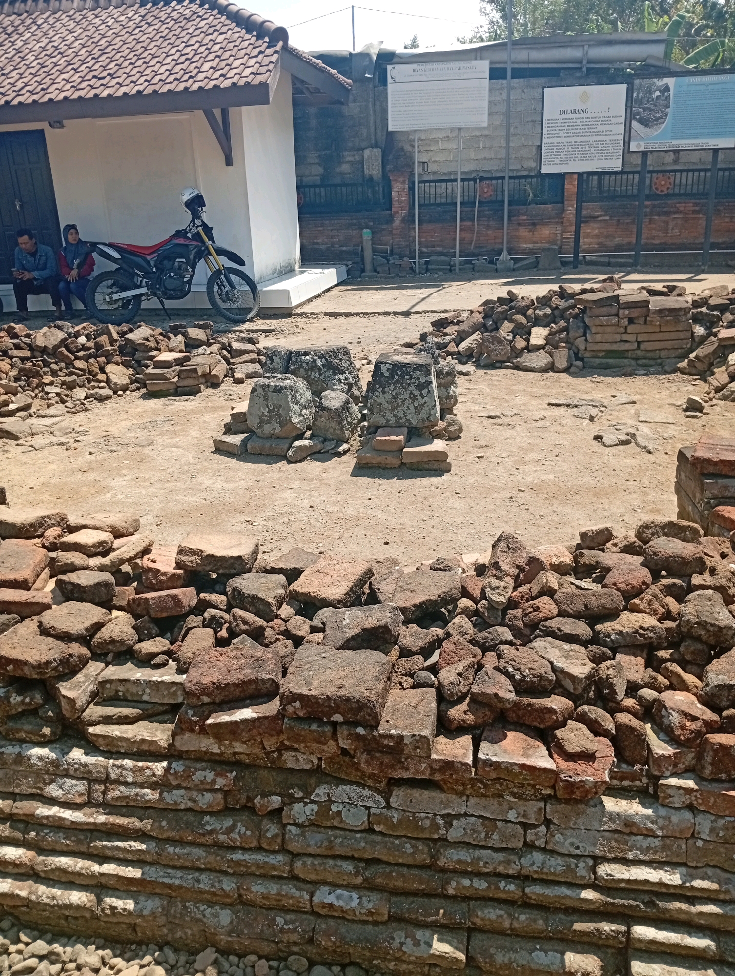 The image depicts an outdoor archaeological site in Kendalbulur, Indonesia. In the foreground, a low, weathered brick wall, possibly part of an ancient structure, is visible, with loose bricks and stones piled on top. Behind this wall, the ground is sandy and uneven, littered with more scattered bricks and stone fragments, suggesting ongoing excavation or preservation work.
Dominating the mid-ground are several piles of bricks and stones, arranged in stacks and loose formations, alongside larger, weathered stone blocks, some of which appear to be carved or shaped, hinting at the remnants of ancient buildings or artifacts.
In the background, a small, white modern building with a terracotta tiled roof stands to the left. Parked beside this building is a red and black dirt bike with a white helmet resting on its handlebars. Two individuals are seated on a low wall or ledge to the left of the building, one of whom is looking at a mobile phone. To the right of the building, a concrete wall supports several informative signs mounted on posts, detailing historical information or regulations, and behind this, a brick fence encloses further structures and trees are visible. The overall environment suggests a historical site undergoing maintenance or interpretation.