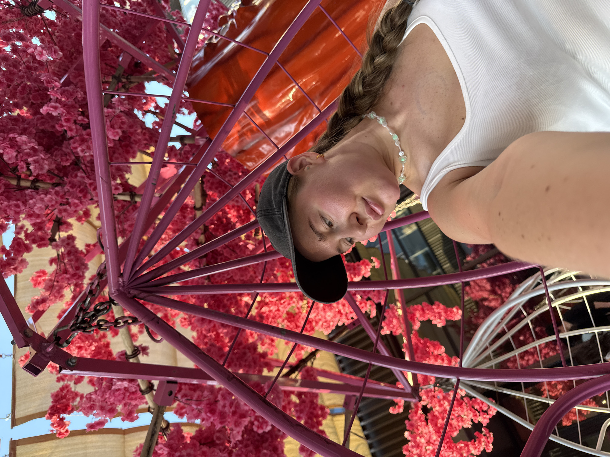 A young Caucasian woman with a braid and a black baseball cap is taking a selfie in Yiwu, China. She is wearing a white tank top and a beaded necklace. She is positioned in front of a large, pink, metallic structure decorated with artificial pink cherry blossoms. Behind her, a bright orange, curved surface is visible. The overall scene suggests an outdoor market or decorative installation, possibly at a shopping center or tourist attraction in Yiwu.