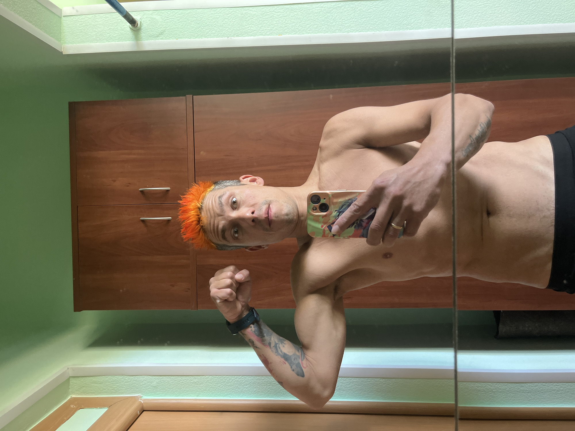A man with orange hair stands shirtless in a room with light green walls and brown cabinets. He is holding a smartphone in his right hand, taking a selfie. He has a watch on his left wrist and tattoos on his left arm. A mirror is visible on the right side of the frame. There is a door and a pipe along the ceiling. The location is possibly Chaikovsky, Russia.