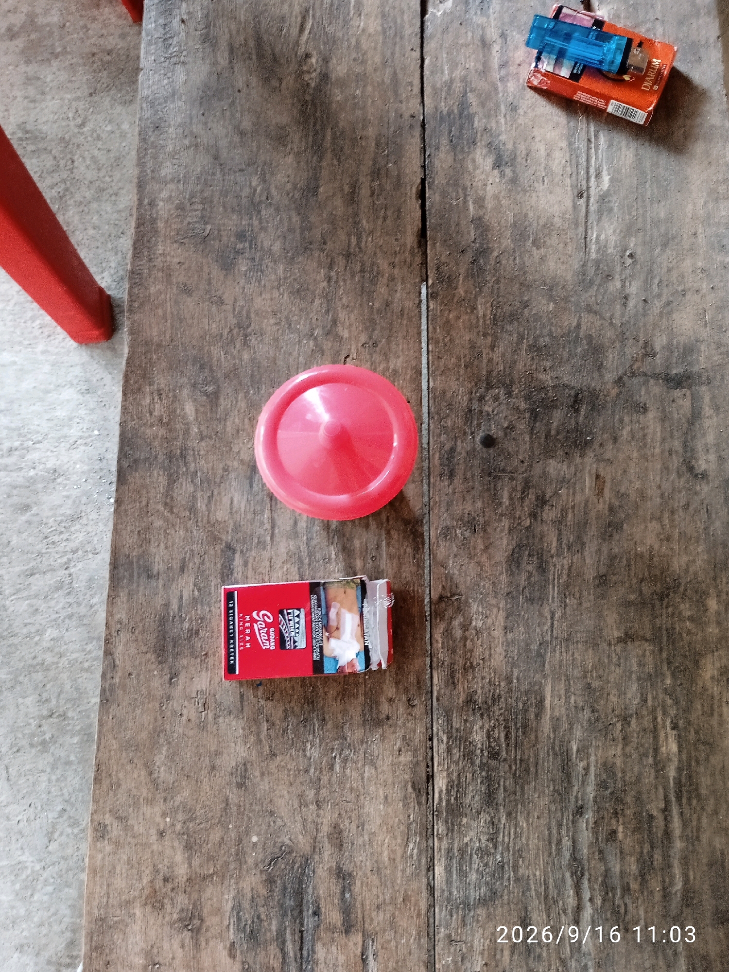 A pack of GUDANG GARAM MERAH KING SIZE cigarettes and a red plastic lid are on a wooden table. A blue disposable lighter rests on a pack of DIARILM cigarettes in the upper right corner. A red chair leg is visible on the left side of the image. The image was taken on September 16, 2026, at 11:03 AM in Kelurahan, Indonesia.