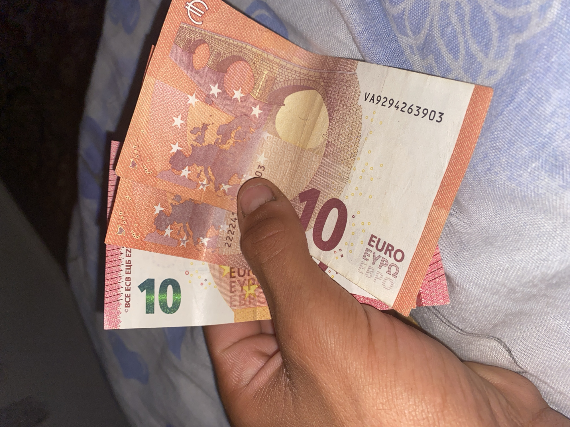 A human hand, with dark skin tone, is holding a fan of euro banknotes. The banknotes appear to be 10 euro bills, as indicated by the large "10" printed on one of them. The bills are predominantly white and orange, with visible security features such as the European flag stars and a watermark. The background consists of a light-colored, patterned fabric, likely a bedsheet or blanket. No specific location in the city of Brashlen, Bulgaria, can be identified from the image.