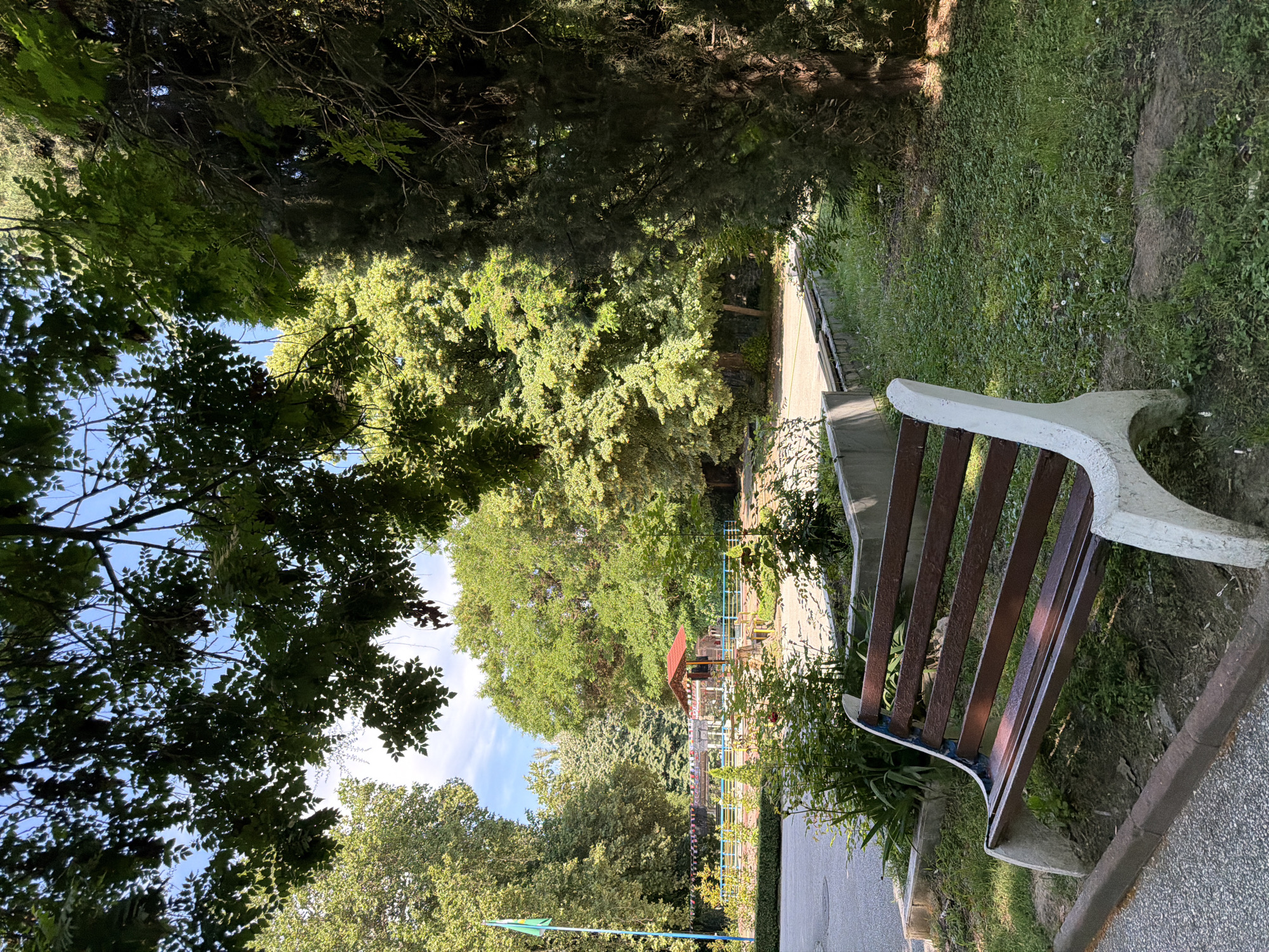 The image depicts a park scene in Pazardzhik, Bulgaria, under a bright, partly cloudy sky. In the foreground, a weathered wooden bench with white concrete supports sits on a patch of grass next to a paved walkway. Behind the bench, a low concrete planter box filled with green foliage separates it from a wider grassy area. In the middle ground, a small, red-roofed pavilion or structure is visible, surrounded by a fence. Beyond this, a grassy area is dotted with trees of various sizes and types, with a flagpole flying a green flag on the left side of the frame. The overall impression is of a public park, likely a place for relaxation and recreation, with lush greenery and clear skies.