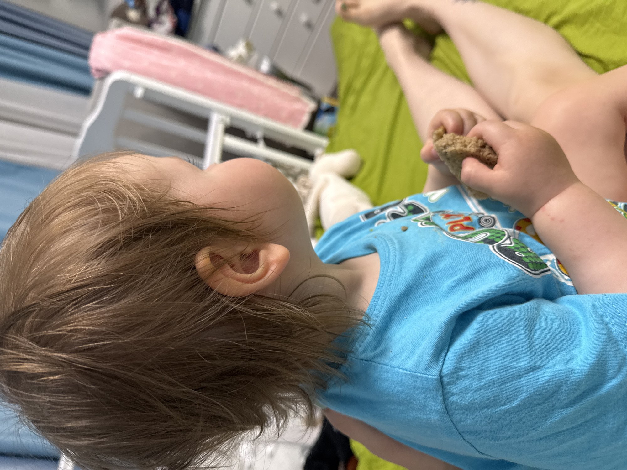 A toddler with light brown hair is lying on a green surface, possibly a bed or blanket, holding a piece of dark bread in their hands. The child is wearing a blue t-shirt with a cartoon graphic. An adult's legs are visible in the background, indicating a parent or caregiver is present. The background includes a changing table with a pink pad, a white dresser, and a blue curtain. The location is impossible to discern from the given image.