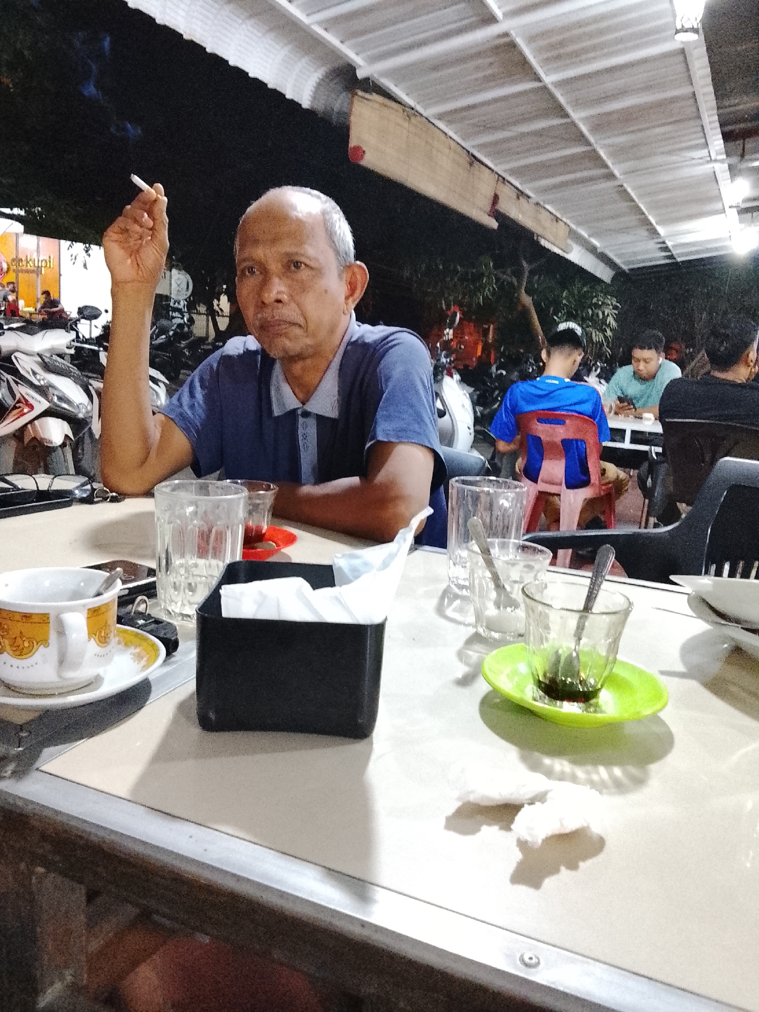 An adult male is smoking a cigarette while sitting at a table in an outdoor cafe. He wears a blue polo shirt. The table in front of him has multiple glasses, a cup of coffee, a black tissue box, and some debris. Other people are seated at tables in the background, one wearing a blue shirt with the number "7" on the back. Scooters are parked behind the cafe area. The cafe has a roof structure made of rectangular panels and is illuminated by lights. This image was taken in Lhokseumawe, Indonesia.