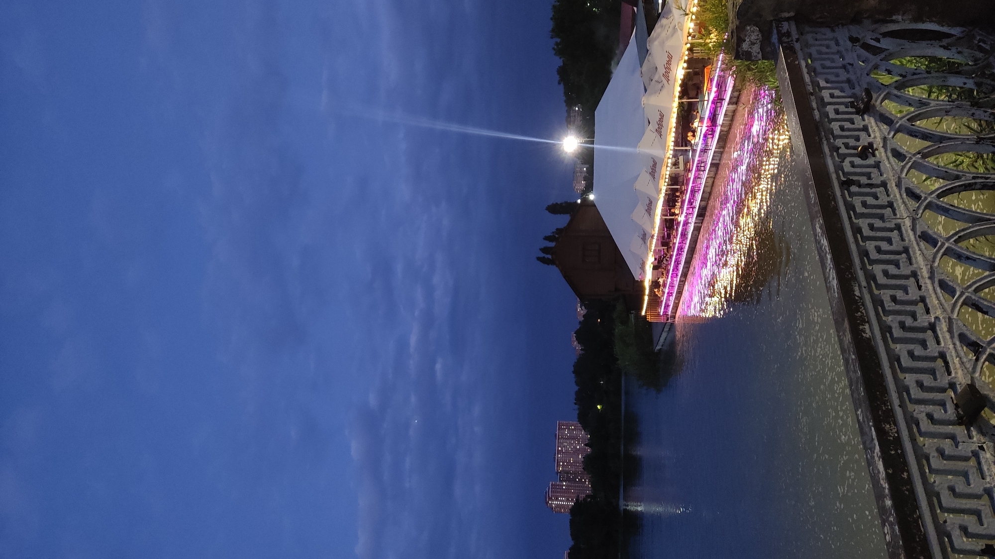 A view of a restaurant on a pier at dusk with a body of water in the foreground and city buildings in the background. The restaurant has a white tented roof with purple and yellow lights illuminating the water beneath it. The water reflects the lights and the dark sky, which has some scattered clouds. In the background, there are trees and two tall buildings with illuminated windows. The scene appears to be from a waterfront promenade with a decorative metal railing in the foreground. The image was taken in Krasnodar, Russia.