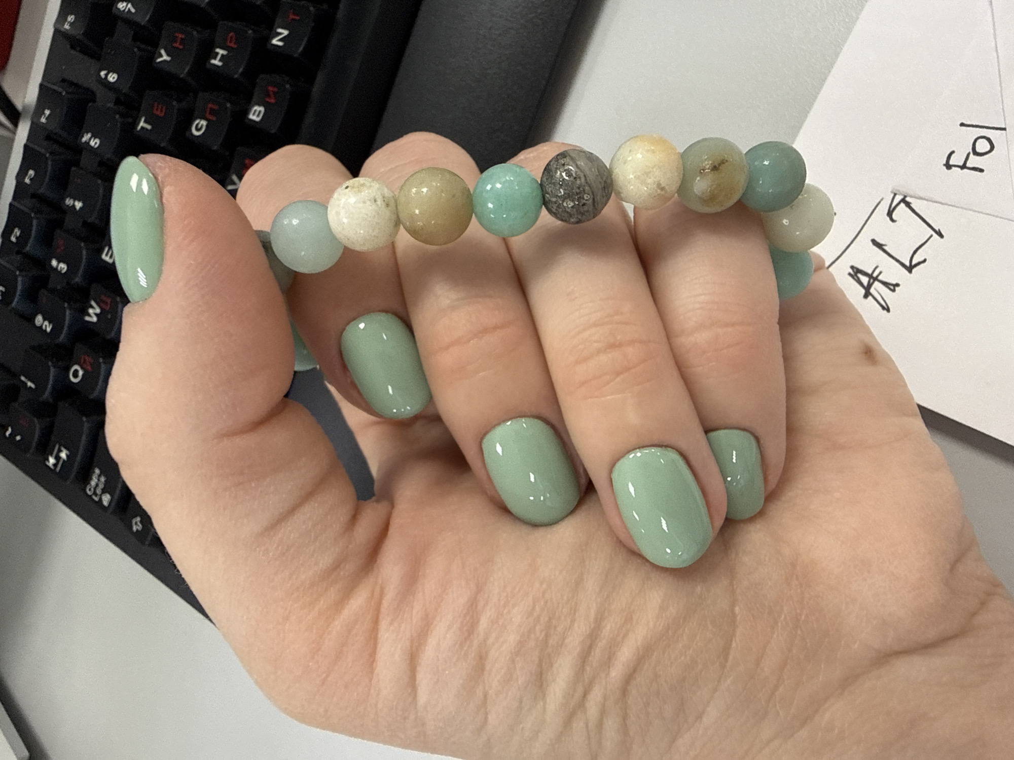 A close-up shot depicts a person's hand adorned with a bead bracelet in varying shades of green, white, and grey. The fingernails are painted in a matching shade of green. A black computer keyboard is visible in the background to the left, along with a piece of paper with handwriting to the right. The words "ALT" and "Fol" are discernible on the paper. The scene appears to be set indoors. There is no evidence to identify a city or country.