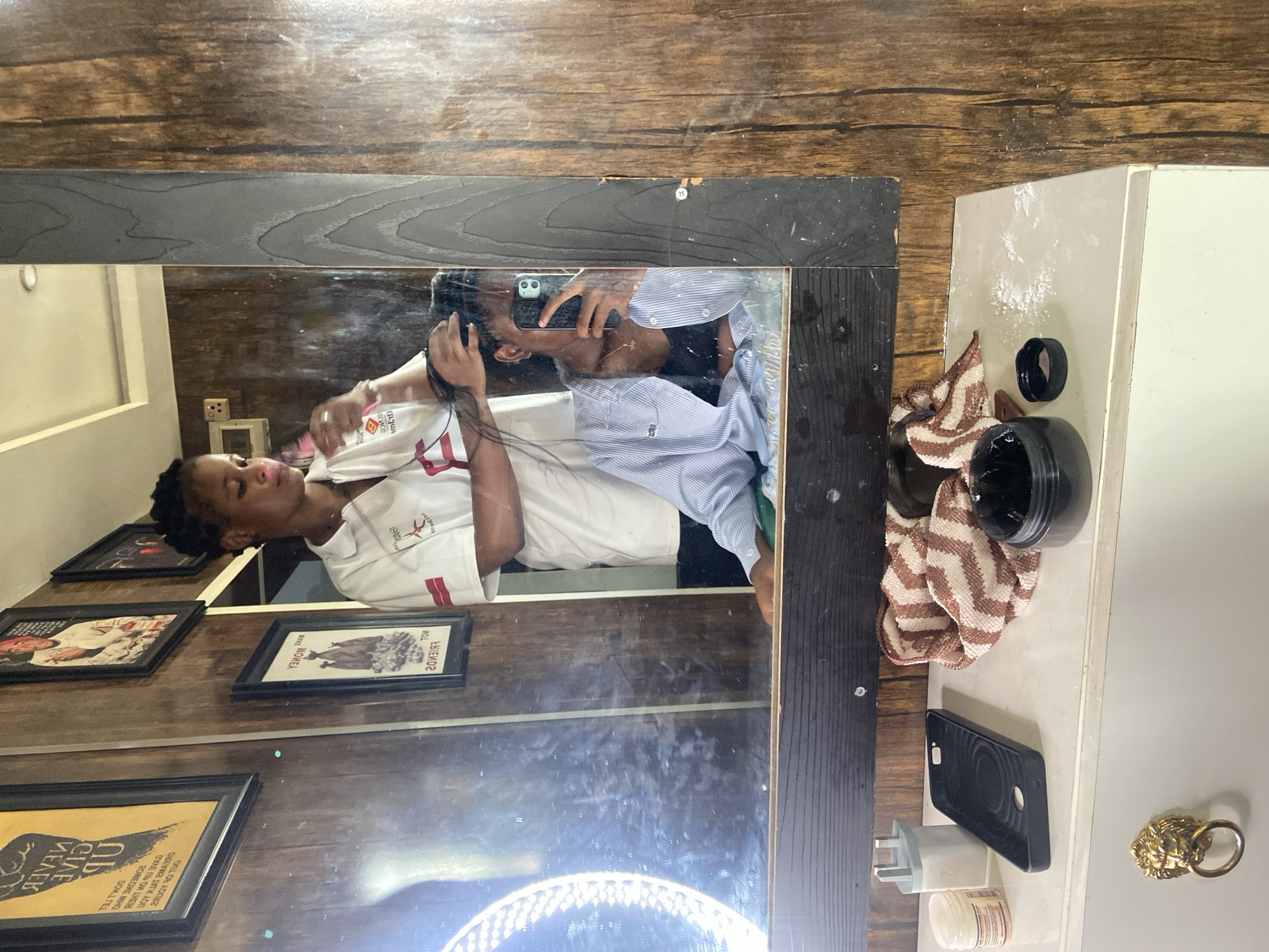 The image is a reflection of a man taking a selfie in a mirror. The man is wearing a white shirt with red accents, and he's holding a smartphone to capture the image. The mirror is framed with dark wood, and behind the man in the reflection, there are framed pictures on a wall. To the right of the mirror, on a countertop, there are various items, including a dark container with a lid, a striped towel, a black phone case, and a charger. The setting appears to be indoors, possibly a barber shop or a personal care space given the presence of what looks like hair product and the general aesthetic. The image does not provide enough information to pinpoint a specific location in Lokoja, Nigeria.