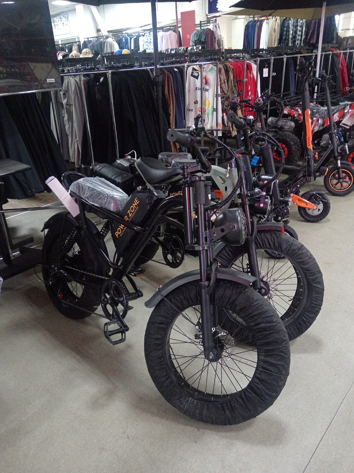 The image shows an indoor retail environment with clothing racks filled with apparel in the background. In the foreground, there are several electric scooters and bicycles displayed. One electric bicycle, a "POWER ZONE" model, is prominently featured in the center. It is black with wide tires that have protective black covers. The bicycle has a black frame, seat, and handlebars, and an orange battery pack. To the right of this bicycle, another electric scooter, black and orange, is visible. A third scooter, also black and orange, is partially visible in the far right background. The floor appears to be a light-colored tiled surface, and shadows are cast from the objects. The overall impression is a retail display area for electric vehicles and possibly clothing. The location is identified as Belogorsk, Russia.