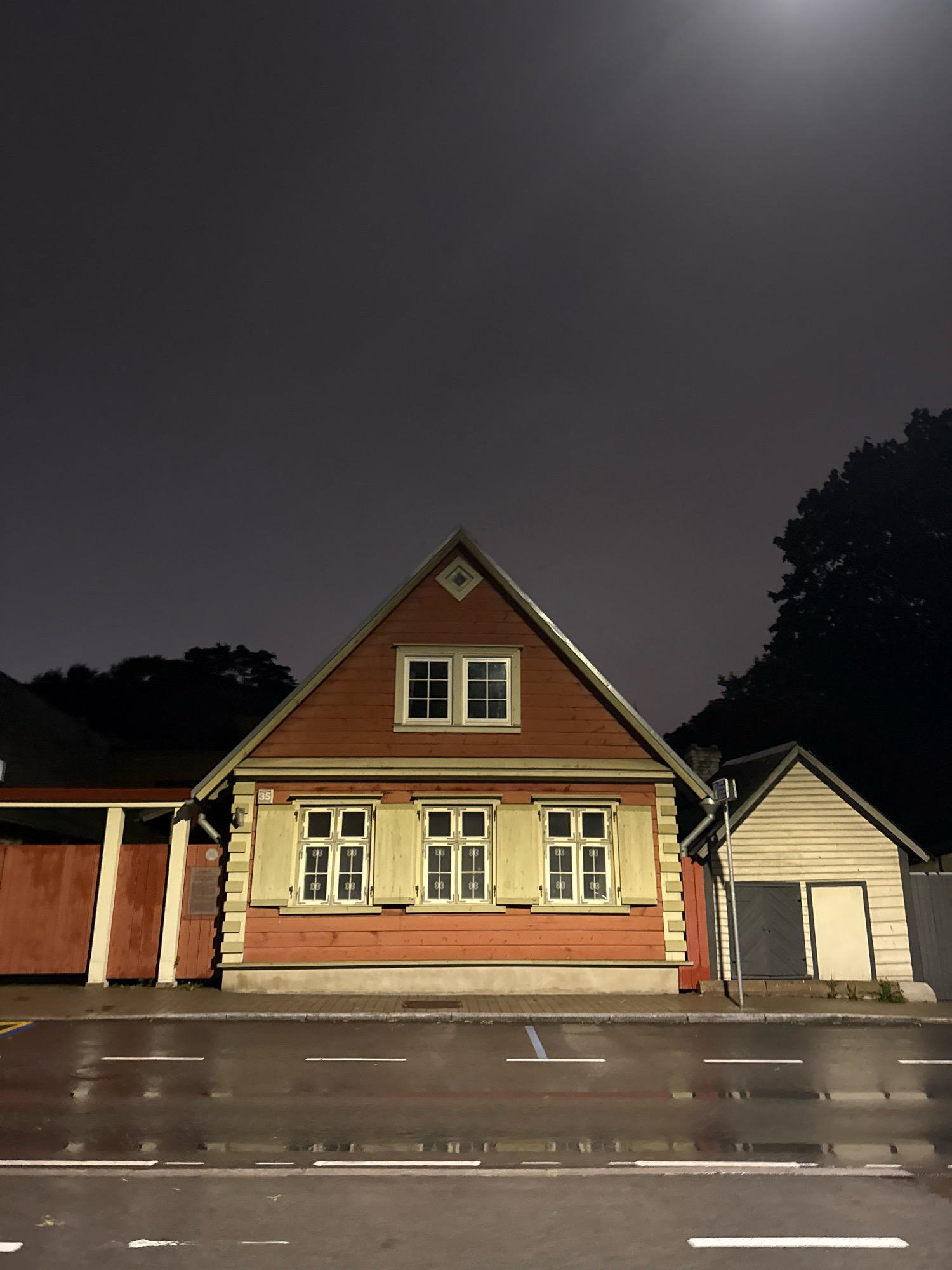 This is a nighttime, eye-level, full-shot photograph of a street in Tallinn, Estonia, likely taken in the Old Town. The sky is dark and overcast with a visible full moon or bright light source in the upper right quadrant, illuminating the scene with a dim, atmospheric glow.
The primary subject is a red, two-story wooden house with a gabled roof. The house has light yellow shutters on its windows, which are illuminated from within, suggesting occupancy. A smaller, light-colored wooden structure, possibly an outbuilding or shed, is attached to the right side of the house. This structure features a dark gray double door and a single lighter-colored door.
In front of the buildings is a wet asphalt road with white lane markings. The wetness of the road indicates recent rain or damp conditions, reflecting the faint light from the sky and buildings. A sidewalk or paved area runs between the road and the buildings. Dark trees are visible behind the buildings, silhouetted against the night sky. The overall atmosphere is quiet and still, evoking a sense of calm and perhaps a touch of mystery due to the low light conditions and the old architecture.