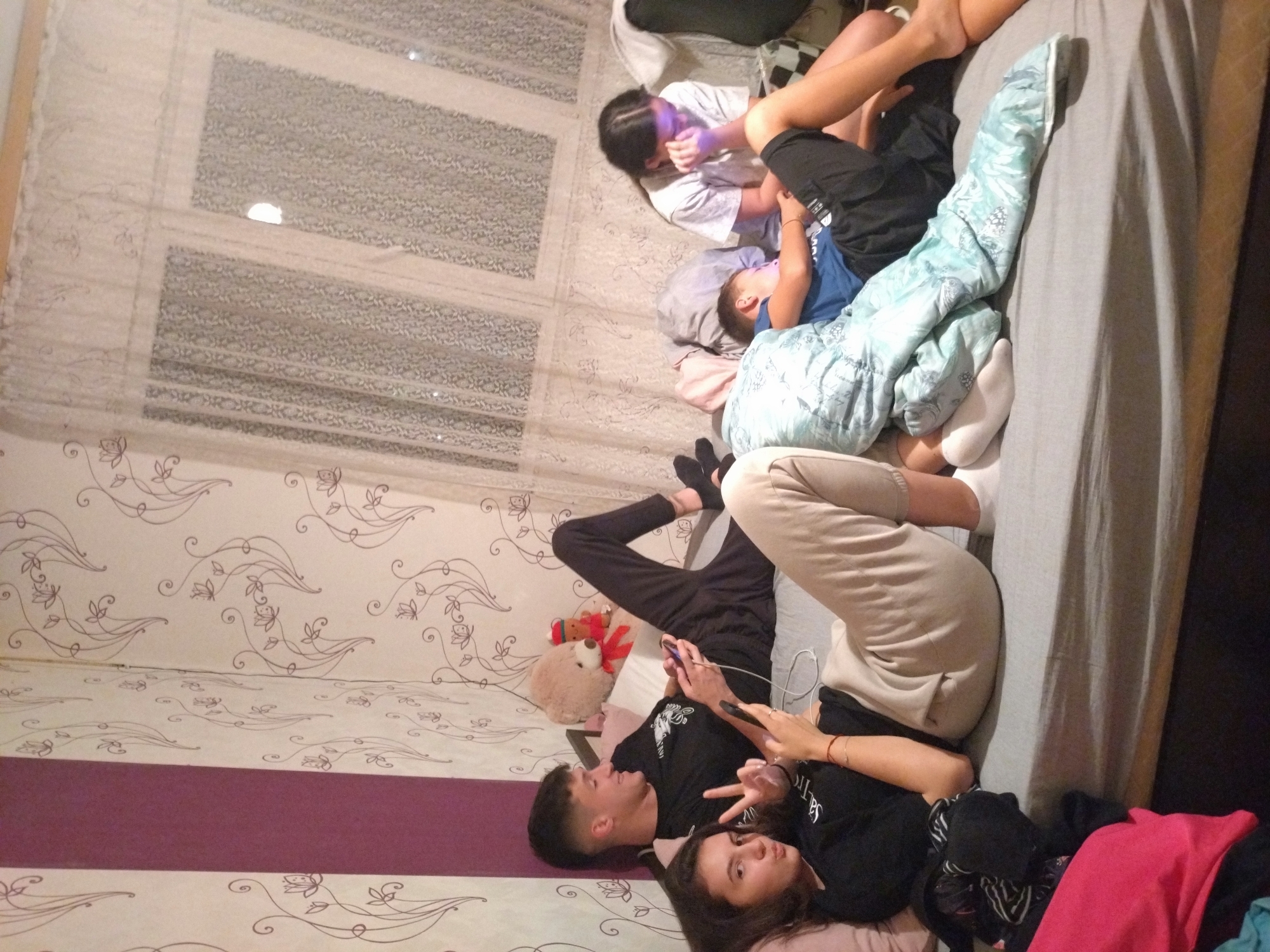 The image shows a bedroom scene, with several individuals present. The room has patterned wallpaper and a window with blinds. The bed is the central element, with multiple people lying on it. One person is holding a mobile phone, and another is making a peace sign. Some individuals appear to be younger, possibly children. A teddy bear is visible near the head of the bed. The location is unspecified and there is no information linking the image to the city of Stara Zagora, Bulgaria.
