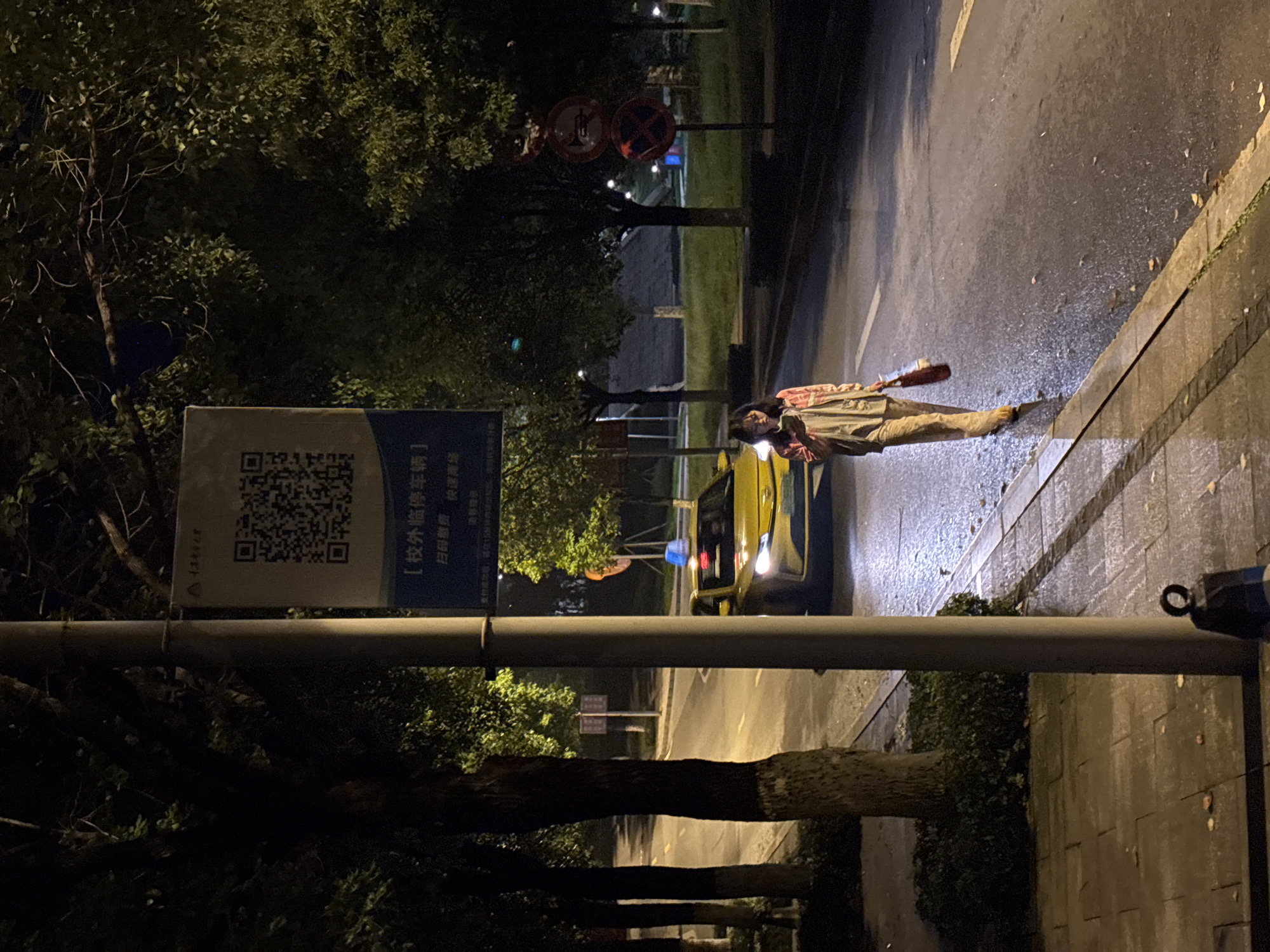 The image shows an urban scene at night in the Nan'an District of China. A person, wearing a plaid shirt and tan pants, lies prone on the wet pavement near a curb. A yellow vehicle with headlights illuminated is adjacent to the person on the road. A sign with a QR code is attached to a utility pole along the sidewalk. Trees are visible in the background. In the distance, traffic signs are present. The environment appears to be wet, possibly after rain. The exact actions or interactions are not clear from the image alone.
