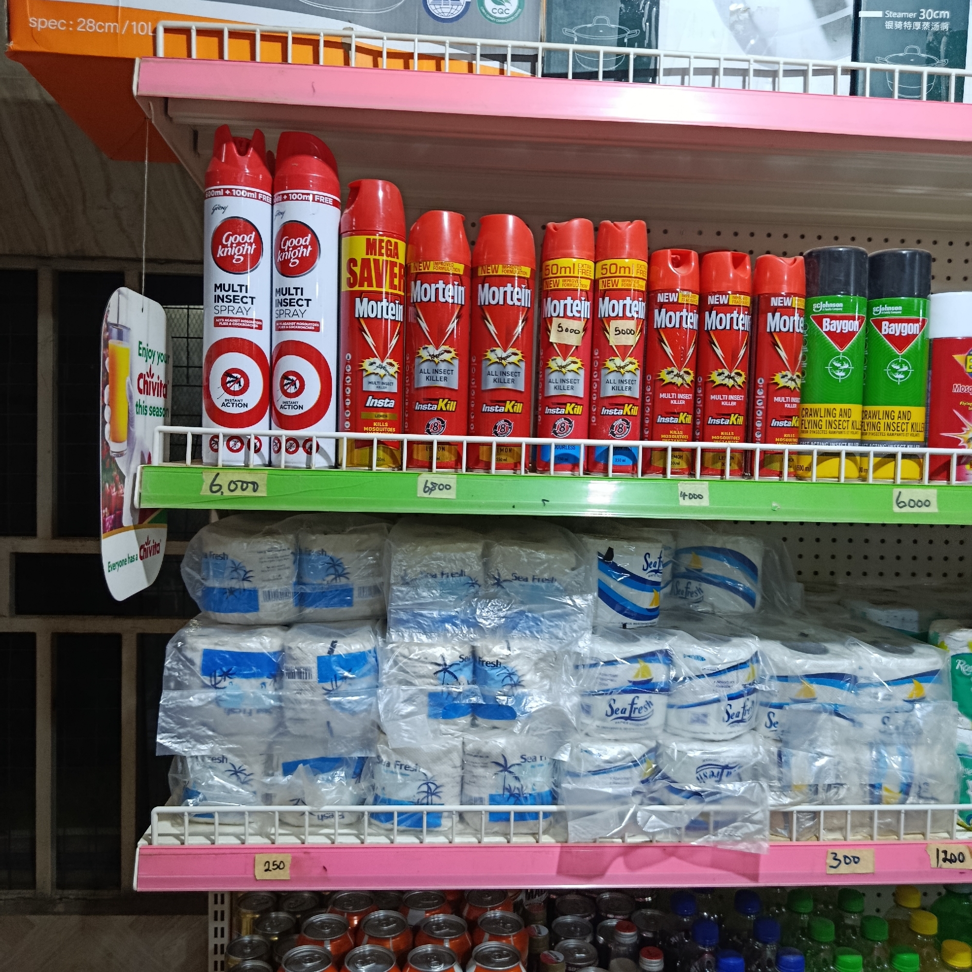 The image displays a well-stocked retail shelf inside a store located in Gwarinpa, Nigeria. The scene is devoid of people.
The uppermost shelf in the background, partially visible, contains packaged items, possibly kitchen appliances, indicated by text such as "Steamer 30cm" and "spec: 28cm / 10L."
The primary focus is the middle shelf, which is divided into two layers. The upper layer showcases various aerosol insect sprays. From left to right, there are two red and white "Good Knight Multi Insect Spray" cans, labeled as "600ml + 100ml FREE" and featuring an "Instant Action" mosquito logo. Adjacent to these are multiple red "Mortein" cans, with various labels including "Mega Saver," "New Improved Formulation," "All Insect Killer," and "InstaKill." Some Mortein cans have handwritten price tags of "5000." Further to the right are two green and black "Baygon Crawling and Flying Insect Killer" cans by SC Johnson. Price tags placed directly below these sprays read "6,000," "6,500," "4,000," and "6,000."
The lower layer of the middle shelf contains numerous individually plastic-wrapped packages of "Sea Fresh" brand paper products, likely toilet paper or paper towels, distinguished by white packaging and blue decorative elements. Below this section, price tags of "250," "300," and "1,200" are visible.
The bottom shelf, partially visible at the very bottom of the frame, holds rows of aluminum beverage cans, predominantly with orange/red tops, and an assortment of plastic bottles with liquids, capped in various colors like green, blue, yellow, and red.
A promotional cardboard hanger for "Chivita" is suspended on the left, displaying the phrases "Enjoy your Chivita this season" and "Everyone has a Chivita," along with an image of a drink in a glass. The background on the far left reveals a dark, gridded structure, possibly a window with security bars. The shelving units are constructed of white wire with pink shelf dividers.