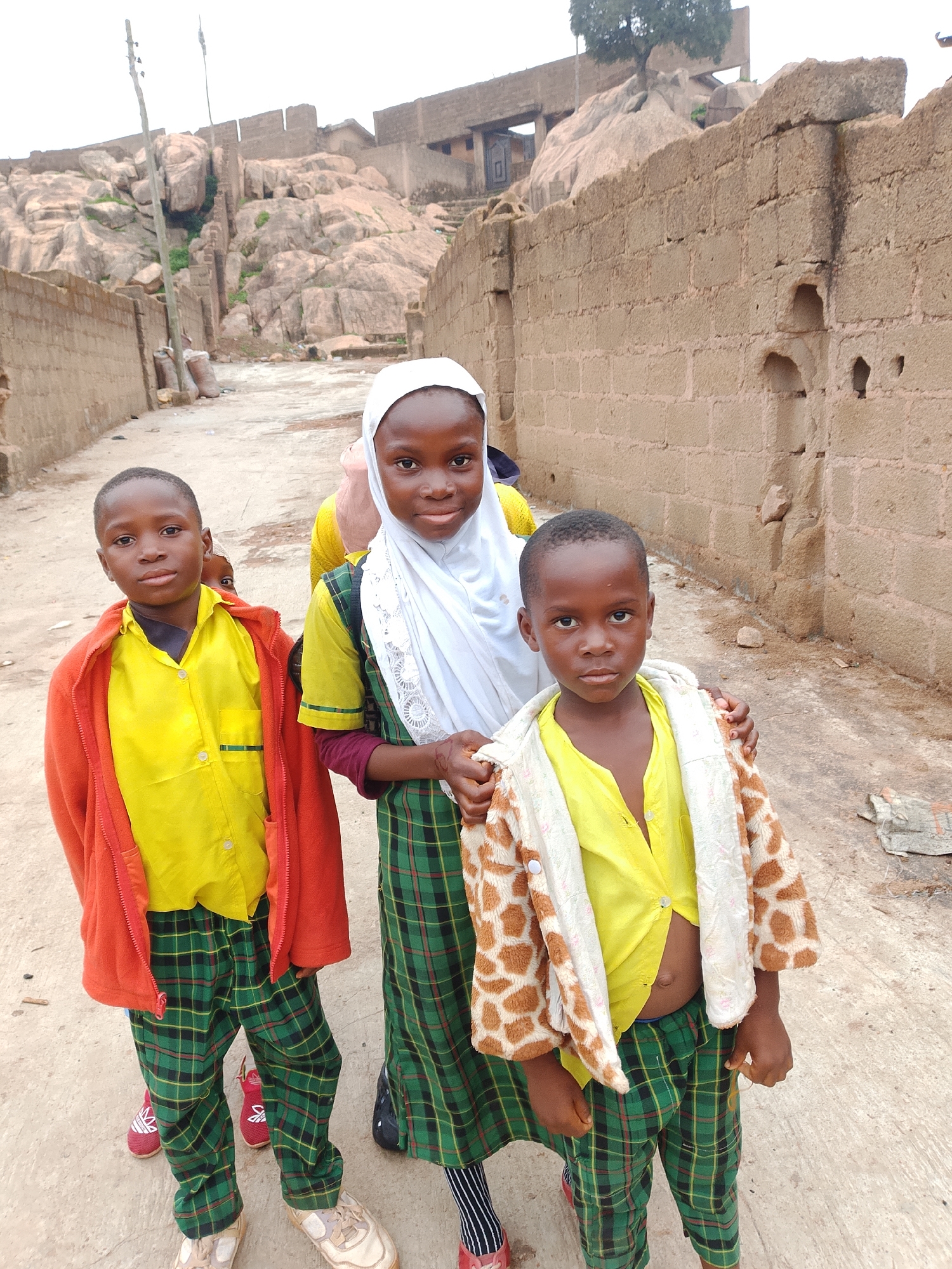 Three children, two boys and a girl, are standing on a dirt path in front of a rough-hewn brick wall and a rocky hillside. The girl, in the center, wears a white hijab and a green plaid dress. The boy on the left wears a red jacket over a yellow shirt and green plaid pants. The boy on the right wears a giraffe-print jacket over a yellow shirt and green plaid pants. The background shows a rocky, uneven terrain with some structures built into the rocks. The sky is overcast.