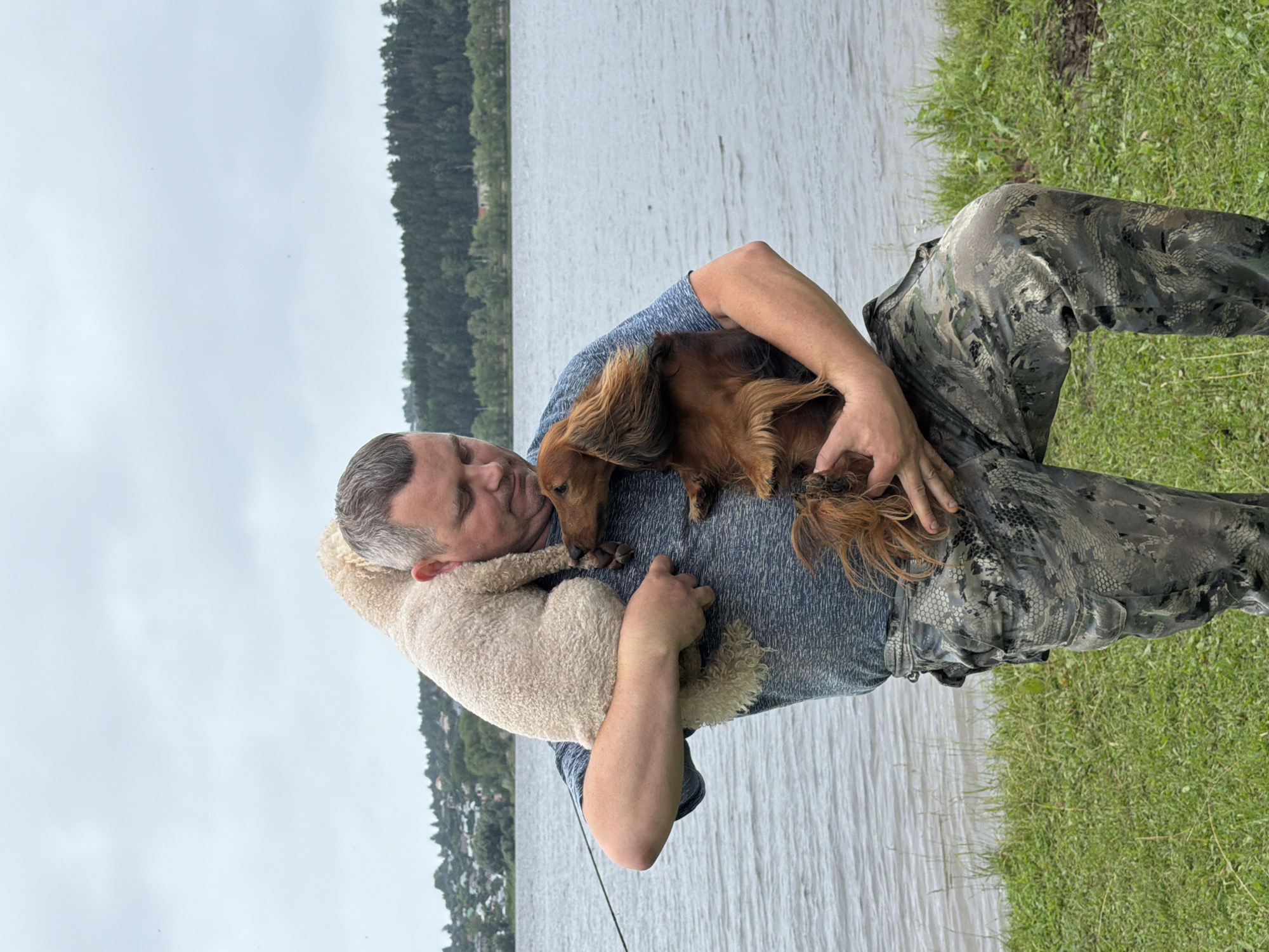 A man, wearing a blue t-shirt and camouflage pants, holds two dogs and a fluffy white animal, possibly a sheep or dog, in his arms. He is standing on a grassy area next to a body of water. In the background, there is a line of trees and a settlement with houses. The sky is overcast. The image appears to be taken on the Kama River embankment in the city of Chaikovsky, Russia.