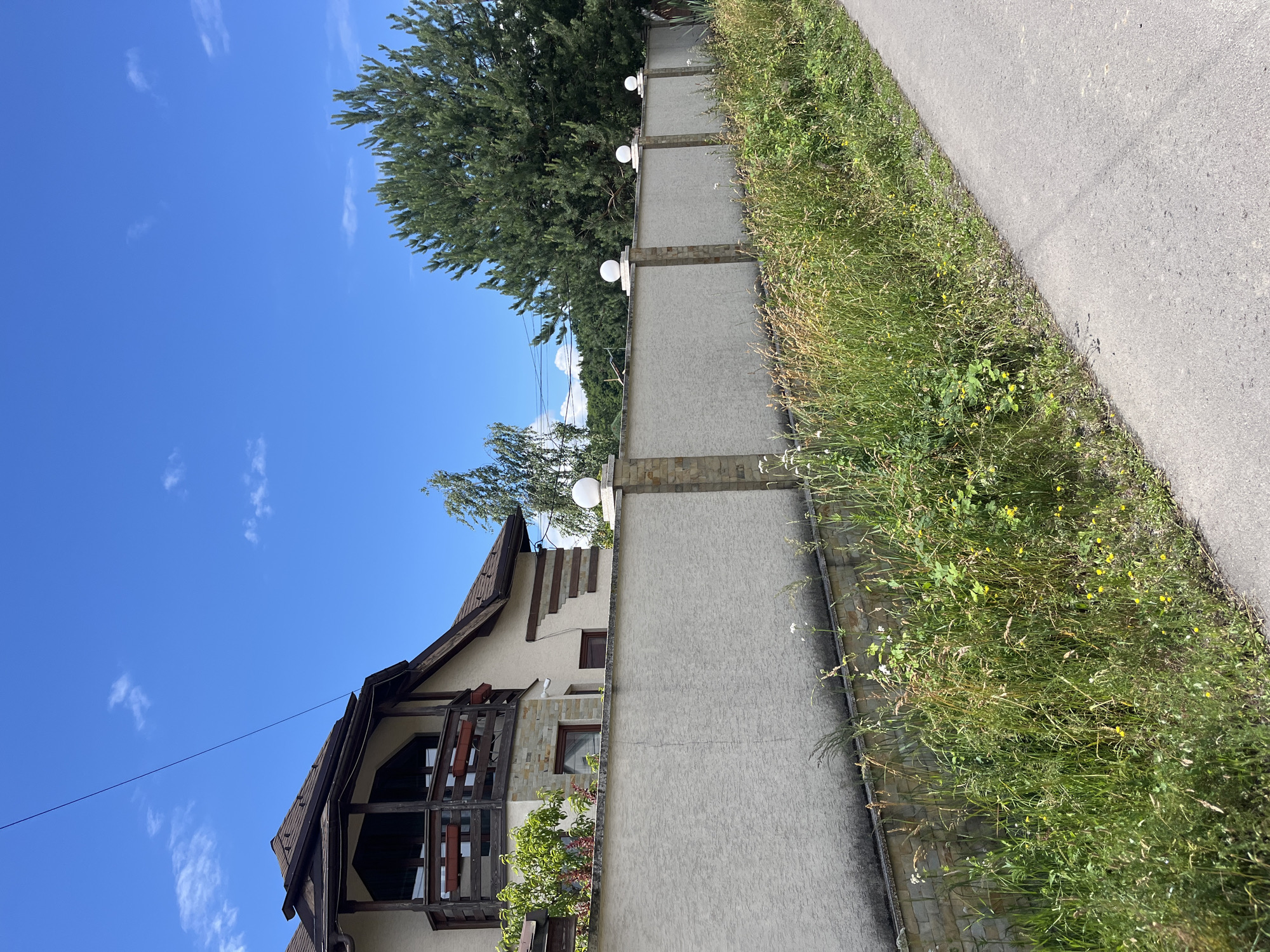 A country house with a brown roof and wooden balcony stands behind a textured beige fence topped with spherical lights. The fence is punctuated by stone pillars. The yard is overgrown with tall grass and wildflowers. A paved road runs alongside the overgrown area. The sky is bright blue with scattered white clouds. The scene is set in the city of Onokivtsi, Ukraine.