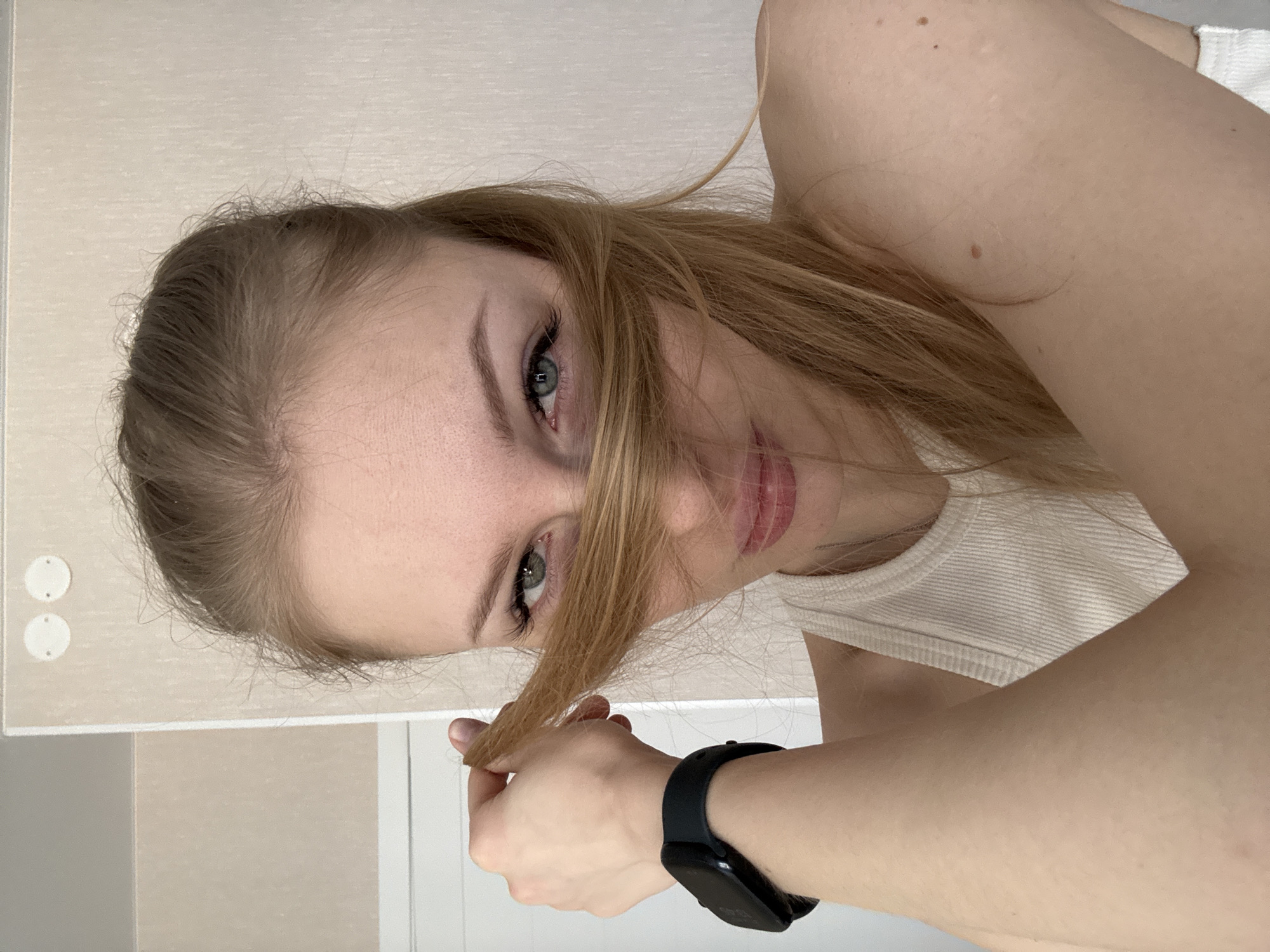 A young woman with light skin and blonde hair is angled, looking directly at the camera. She's holding a strand of her hair in her right hand, which has a black smartwatch on the wrist. She is wearing a white ribbed tank top. The background is a plain, light-colored wall. There are two round, white objects affixed to the wall to the upper left. The location in Perm, Russia cannot be determined from the visual information.
