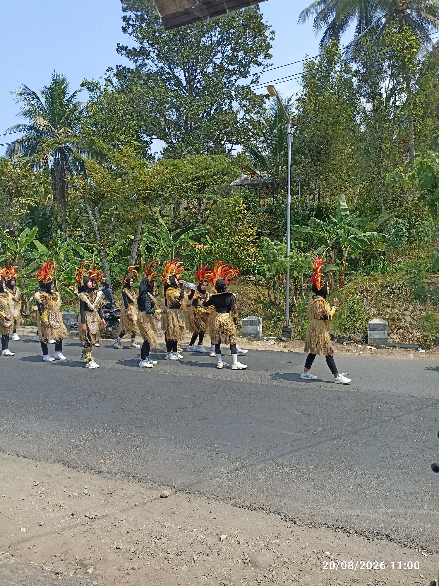 A group of individuals in traditional attire are participating in a parade on a paved road in Munjungan, Indonesia. The participants are adorned in costumes featuring straw skirts, black tops, and elaborate headpieces with red and yellow feathers. They are walking in a line, some with their hands raised as if performing. The background consists of lush green trees and vegetation, typical of a tropical environment, under a clear blue sky. A lamppost and a small shelter are visible on the side of the road, along with some rocks and debris on the unpaved shoulder. The scene suggests a cultural celebration or event taking place outdoors.