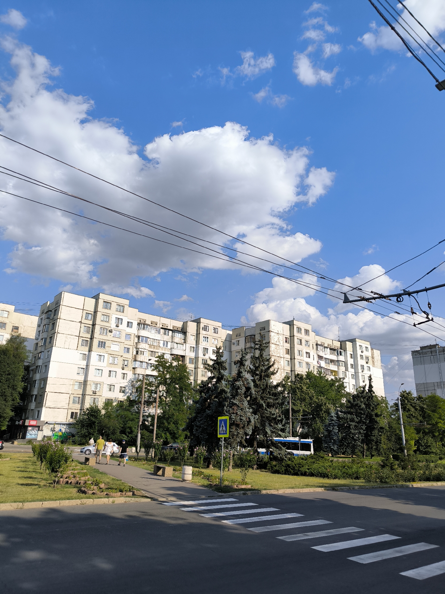A medium shot captures a wide, paved street in Chișinău, Moldova, with a pedestrian crosswalk in the foreground. Beyond the street, a sidewalk borders a grassy area with trees and a large, multi-story apartment building. Overhead, a complex network of trolleybus wires stretches across the frame. The sky is a vibrant blue with fluffy white clouds. On the sidewalk, three people are walking away from the camera, and a silver car is parked nearby. A blue and white trolleybus is visible in the background behind the trees. A yellow pedestrian crossing sign is posted on the left side of the sidewalk. The apartment building, a typical Soviet-era block, has a light-colored facade and numerous windows.