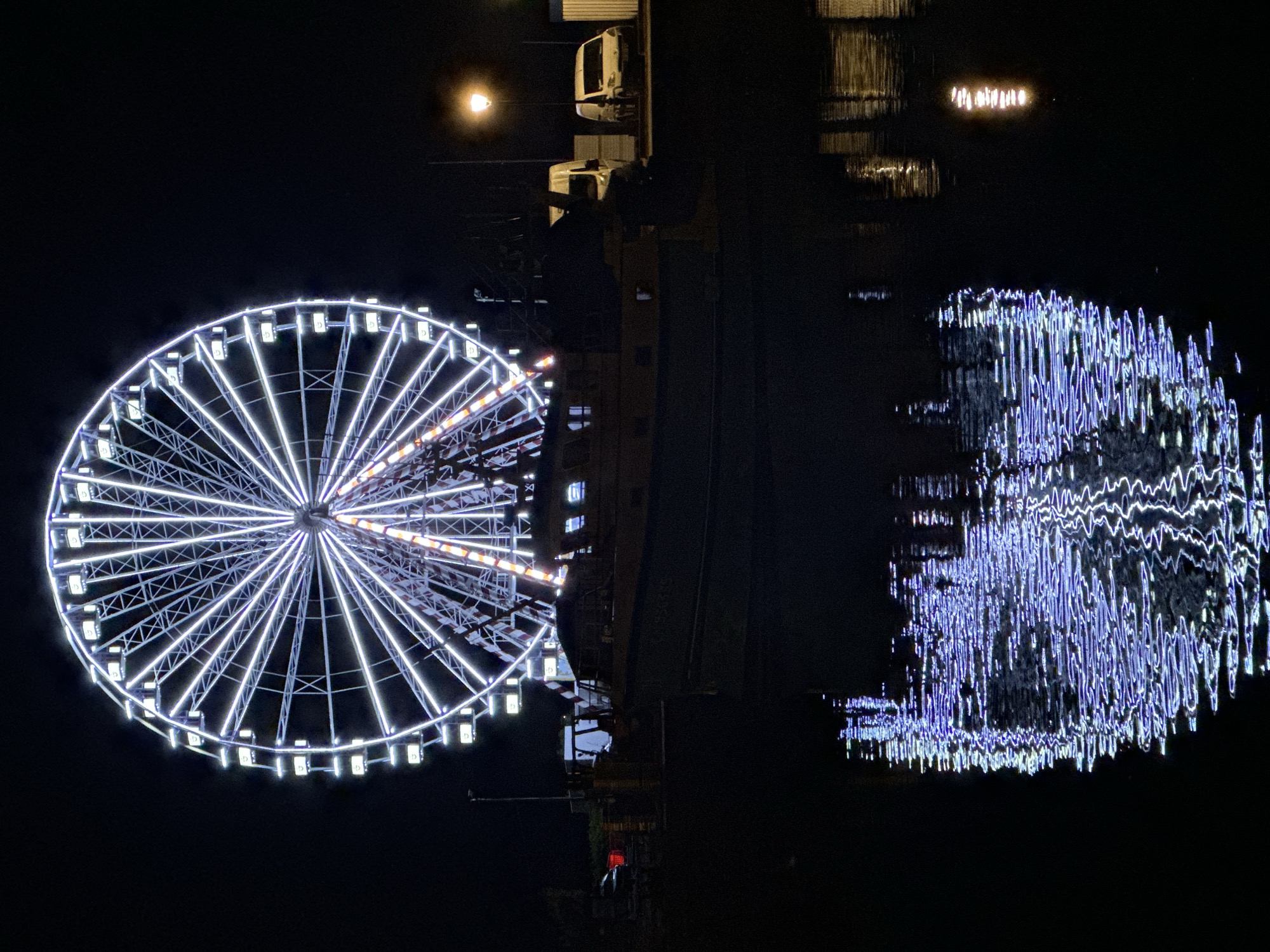 This is a night photograph of a Ferris wheel illuminated with white lights, situated on a waterfront. The Ferris wheel dominates the upper portion of the image, its intricate structure and bright lights casting a glow against the dark sky. Below the Ferris wheel, a boat is docked, its silhouette visible against the water. The lights from the Ferris wheel and other harbor lights are reflected in the dark water, creating a shimmering, abstract pattern of blue and white. In the background, there are some parked vehicles and streetlights, suggesting an urban or harbor setting. The overall scene is one of a lively evening at a seaside town.
The Ferris wheel depicted in the image is located in the city of Honfleur, France, specifically at the Vieux Bassin (Old Harbor). This location is a prominent tourist attraction known for its picturesque harbor, historic architecture, and vibrant atmosphere, especially at night when the lights illuminate the scene.