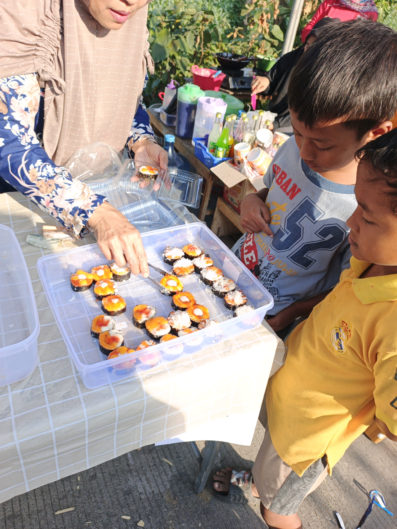 In Jatipasar, Indonesia, on a sunny day, a woman in a floral-patterned blue shirt and a beige headscarf is arranging sushi rolls in a clear plastic container. Her hands, equipped with tweezers, are meticulously placing sushi pieces that have orange toppings and white garnishes into the container. Beside her, on a table covered with a checkered cloth, other sushi rolls are visible. Two young boys stand near the table, observing the sushi. One boy, wearing a blue t-shirt with the number "52" and the word "URBAN" printed on it, leans in towards the sushi. The other boy, in a bright yellow polo shirt, looks on with apparent interest. In the background, various bottles and containers, possibly containing drinks or other food items, are arranged on a table or stall. The setting appears to be an outdoor market or street food stall, with greenery and a glimpse of buildings in the distance. The ground is paved with concrete.