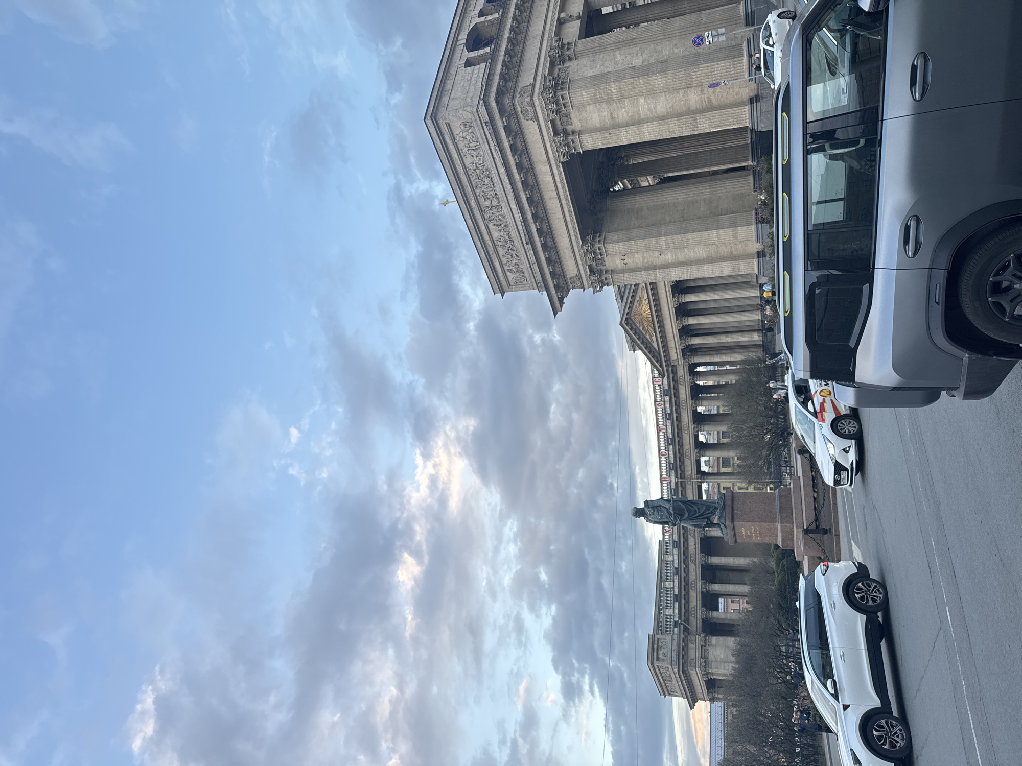 The image displays an urban scene in Saint Petersburg, Russia, centered around the majestic Kazan Cathedral. The sky is partially cloudy with patches of blue, suggesting an overcast day or perhaps late afternoon with soft, diffused light.
The most prominent subject is the Kazan Cathedral, occupying the left side of the frame. Its grand neoclassical facade features an extensive colonnade of large Corinthian columns and ornate friezes along its roofline. In front of the cathedral, a dark bronze statue of a man, likely Field Marshal Mikhail Kutuzov, stands on a stone pedestal, facing towards the viewer.
The foreground and right side of the image feature a city street with vehicles. A modern, dark gray SUV-like car with distinctive yellow accents along its roofline is parked or slowly moving on the far right. Further down the street, a white crossover vehicle is visible, and behind it, another white car bears "SHELL" branding on its side. A circular blue street sign with a red diagonal line, indicating "no stopping" or "no waiting," is partially visible behind the gray car.
Small figures, appearing to be pedestrians, can be seen walking on the street and near the statue, indicating a public space. The scene captures typical daily activity in a busy city environment, combining historical architecture with modern urban elements. The text on the statue's pedestal is not legible.
