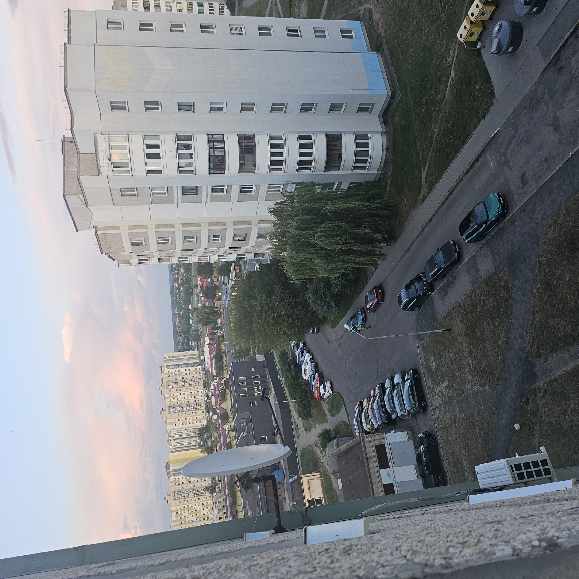 An aerial view of a residential area in Brest, Belarus, depicts several apartment buildings, a satellite dish, and a street lined with parked cars. The sky is filled with soft pink and white clouds, suggesting either sunrise or sunset. The buildings are predominantly light-colored, with multiple stories and numerous windows. A large, white satellite dish is mounted on the side of one building. Cars of various colors are parked along the street and in designated parking areas. Green trees and grass are visible, indicating a well-maintained urban environment. The scene appears peaceful and ordinary, characteristic of a typical residential neighborhood.