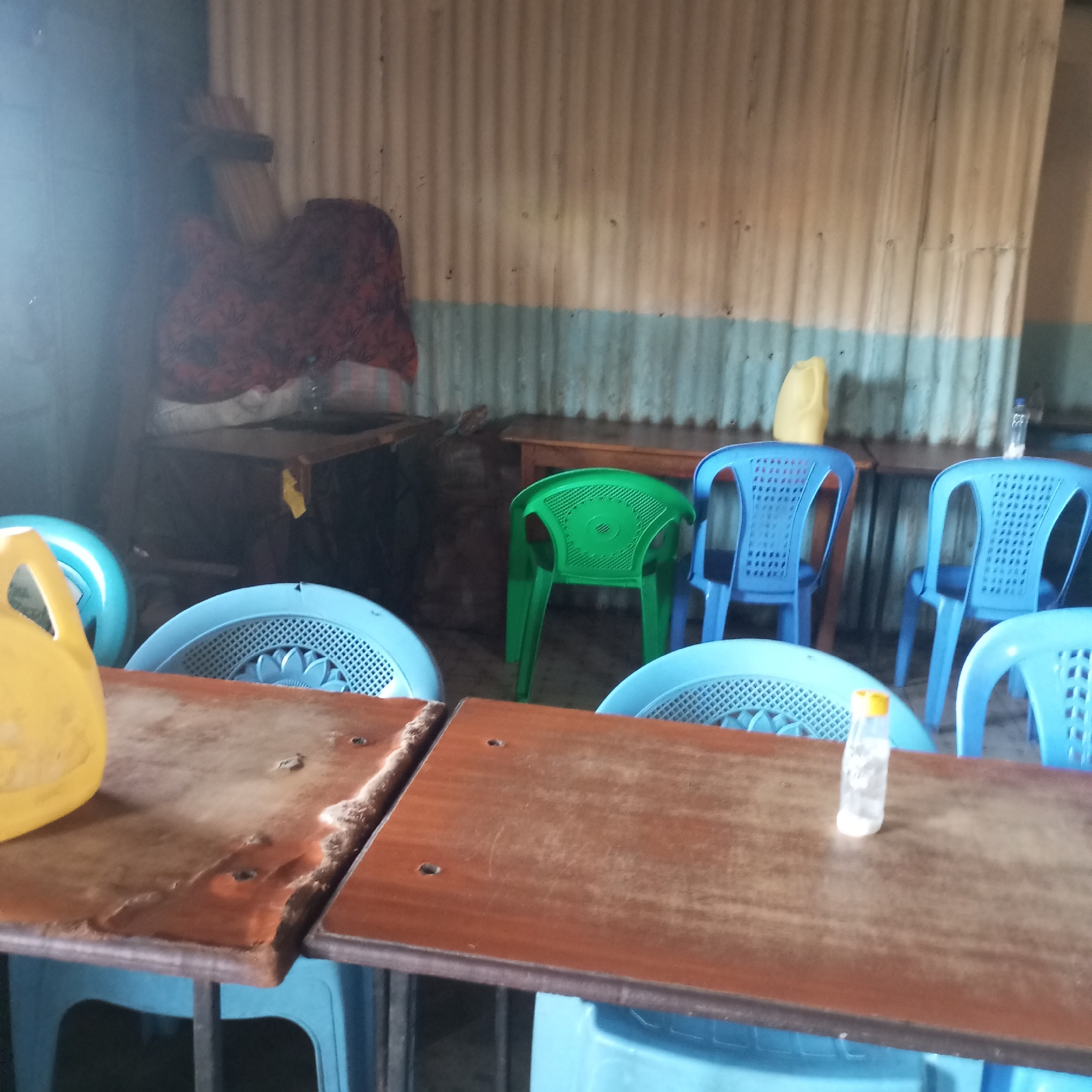 The image shows an interior space in Sheywe ward, Kenya. The room is furnished with several light blue plastic chairs and wooden tables. A green plastic chair is also visible. The walls are made of corrugated metal with a blue stripe at the bottom. Some items, including what appears to be a cloth, are stored on a shelf or surface in the background. There is a yellow plastic container on one table and a small bottle with a yellow lid on another. The tables have a worn appearance.