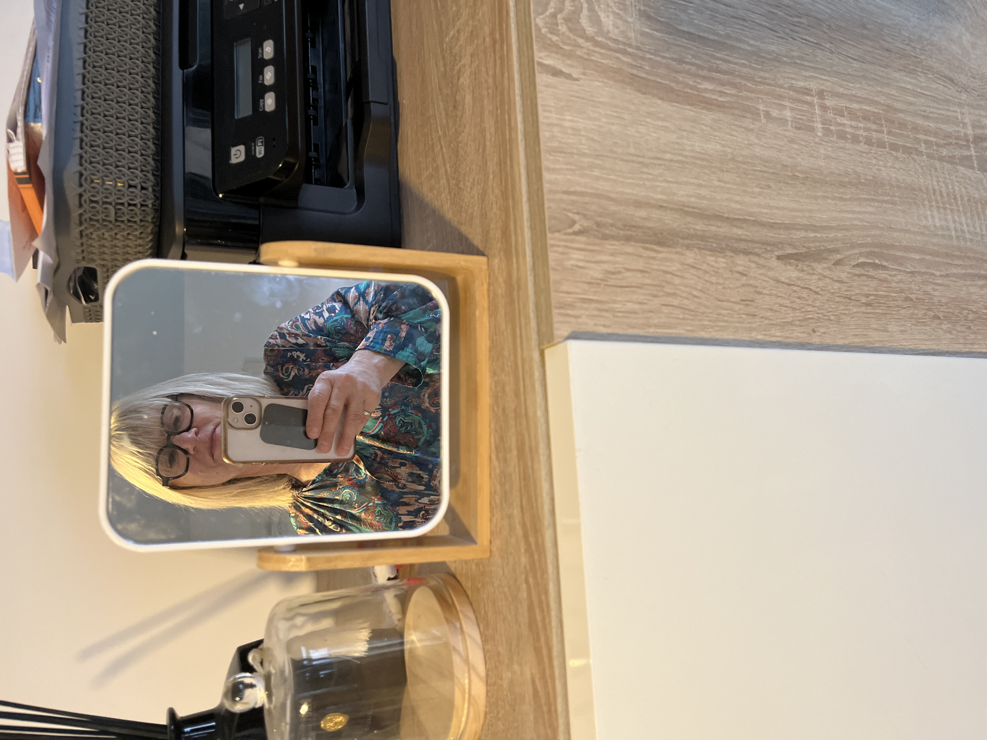 The image depicts an indoor scene in Chambœuf, France. A blonde woman, wearing dark-rimmed glasses and a multicolored patterned blouse, is reflected in a rectangular mirror with rounded corners, framed in light wood. She holds a light-colored smartphone with a dark rectangular attachment on its back in her right hand, positioned as if taking a photograph of her reflection. A ring is visible on her left ring finger.
The mirror rests on a light wood-grain surface. To the left of the mirror, a tall, slender black diffuser bottle with black reeds and a clear glass cloche with a wooden base are present. To the right of the mirror, a black HP multifunction printer is visible, topped by a dark grey woven plastic basket filled with various papers and an orange folder. Below the light wood-grain surface, a white cabinet door is visible on the left, while the wood-grain pattern continues on the right side of the lower portion of the image. The background wall is a plain, light cream color.