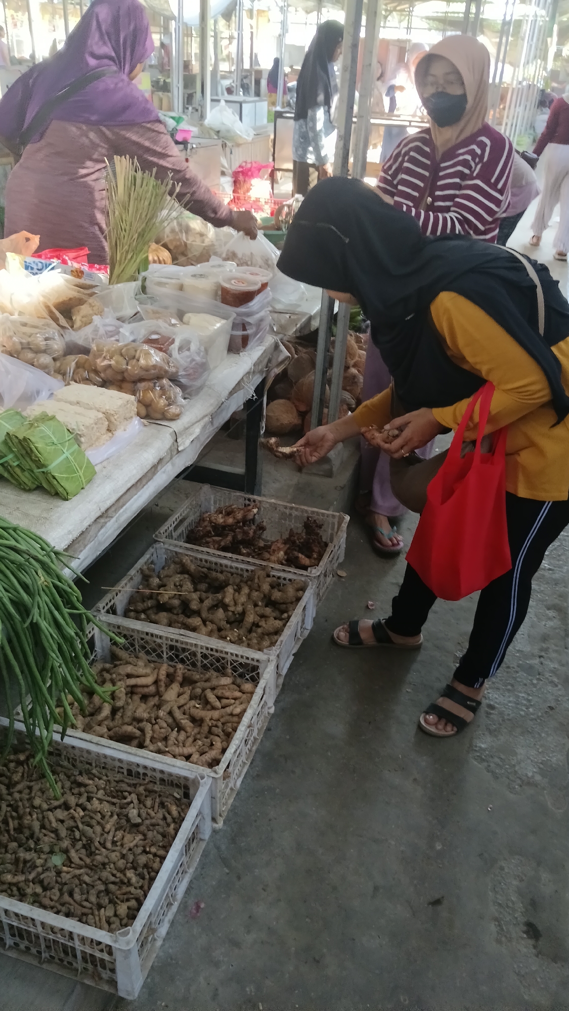 In the city of Dukuh, Indonesia, a marketplace scene unfolds with a focus on produce and local commerce. The foreground is dominated by several white plastic crates filled with various root vegetables, likely ginger or turmeric, arranged in neat rows on the concrete ground. To the left, a bundle of long green beans hangs, adding a splash of vibrant color to the scene. A woman wearing a yellow shirt and black pants with white stripes, and a black hijab, is the central figure, bending over a stall and examining produce. She holds a bright red tote bag in her right hand and is reaching out to select a root vegetable from a display on the stall.
Behind her, another woman in a purple hijab and patterned shirt is also engaged in the market activities, with her hands near a selection of bagged goods. Further back, a woman with a beige hijab and a striped shirt, wearing a face mask, stands near other shoppers and market stalls. The background reveals more of the market's environment, with covered stalls, various goods on display, and other individuals browsing or working. The lighting suggests it is daytime, casting shadows and highlighting the textures of the produce. The overall atmosphere is one of bustling everyday life in an Indonesian market.
