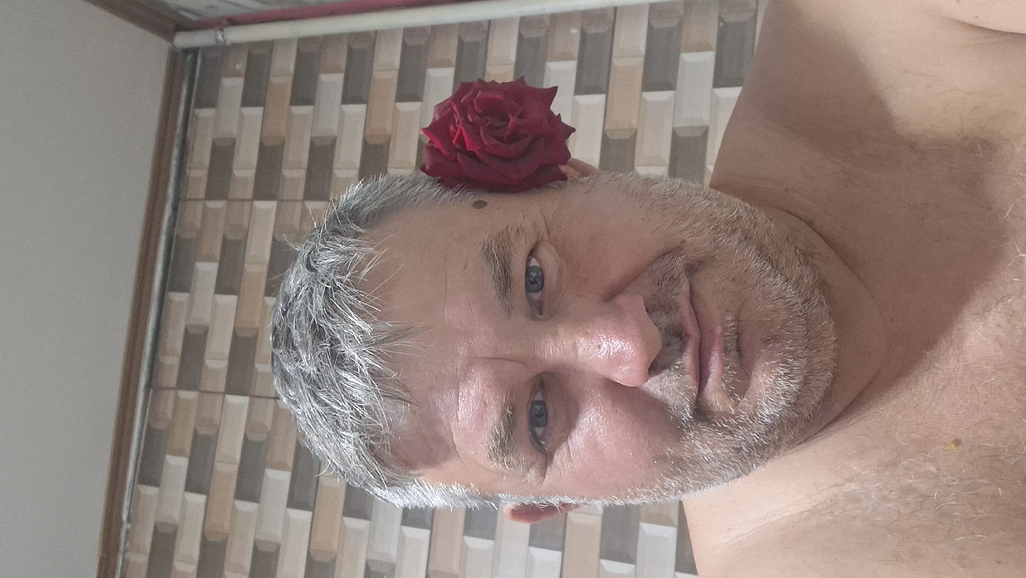 A close-up, eye-level, waist-up shot shows a middle-aged Caucasian man with fair skin, graying hair, and a beard. A red rose is tucked behind his right ear. He has blue eyes and a mole on his right cheek. The background consists of a tiled wall with rectangular tiles in varying shades of beige and brown, and a white ceiling. The lighting is bright, with some shadows visible on the man's face and neck. The image appears to be a selfie, taken indoors, possibly in a bathroom given the tiled background. The man is shirtless, exposing his shoulders and upper chest. His expression is neutral. The overall scene is intimate and personal. The specific location in Voinova, Moldova is not discernible from the image itself.