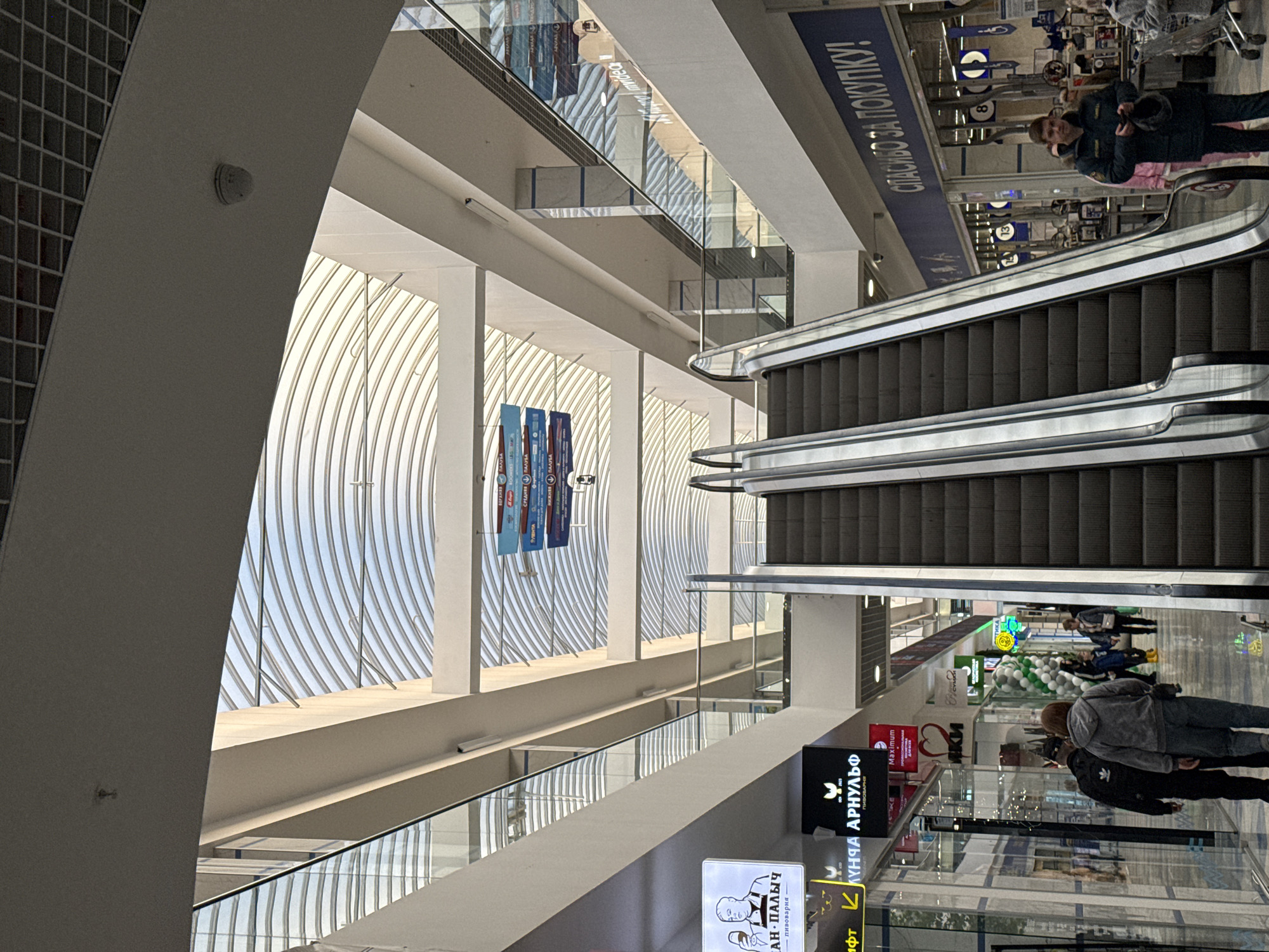 This indoor, eye-level shot, taken in Izhevsk, Russia, features the interior of a shopping mall. The mall is multi-level, evident from the escalator dominating the frame from top right to bottom center. People are walking through the mall; one individual, dressed in what seems like a uniform, can be seen near the top of the escalator. Retail shops, marked with signage in Russian, line the lower levels. The mall is well-lit, with daylight filtering through a curved, ribbed glass ceiling. The design includes modern architectural elements, like exposed columns and glass railings. Signage for brands like "AH-ПАЛЫЧ" and "Maximum" are visible, alongside Russian text like "СПАСИБО ЗА ПОКУПКУ!" (Thanks for the purchase!). The levels include "ПАЛУБА", "СРЕДНЯЯ ПАЛУБА", and "НИЖНЯЯ ПАЛУБА".