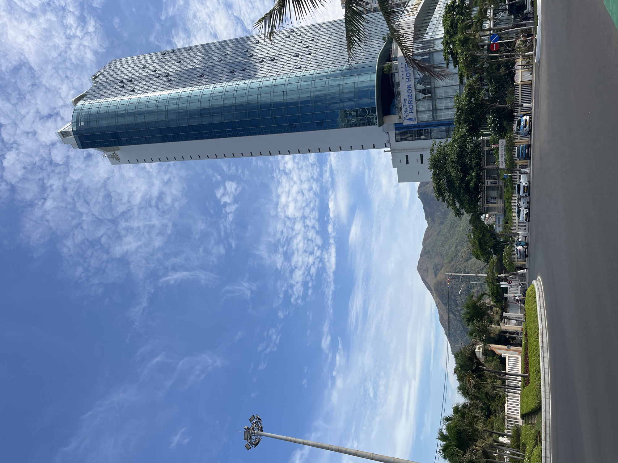 The image shows a view in Nha Trang, Vietnam. A tall glass-fronted building, the "Horizon Hotel", dominates the left side of the image. The sky is blue with scattered white clouds. In the background, there's a view of a mountain. Along the right side of the image, a street is visible with parked cars and vegetation.