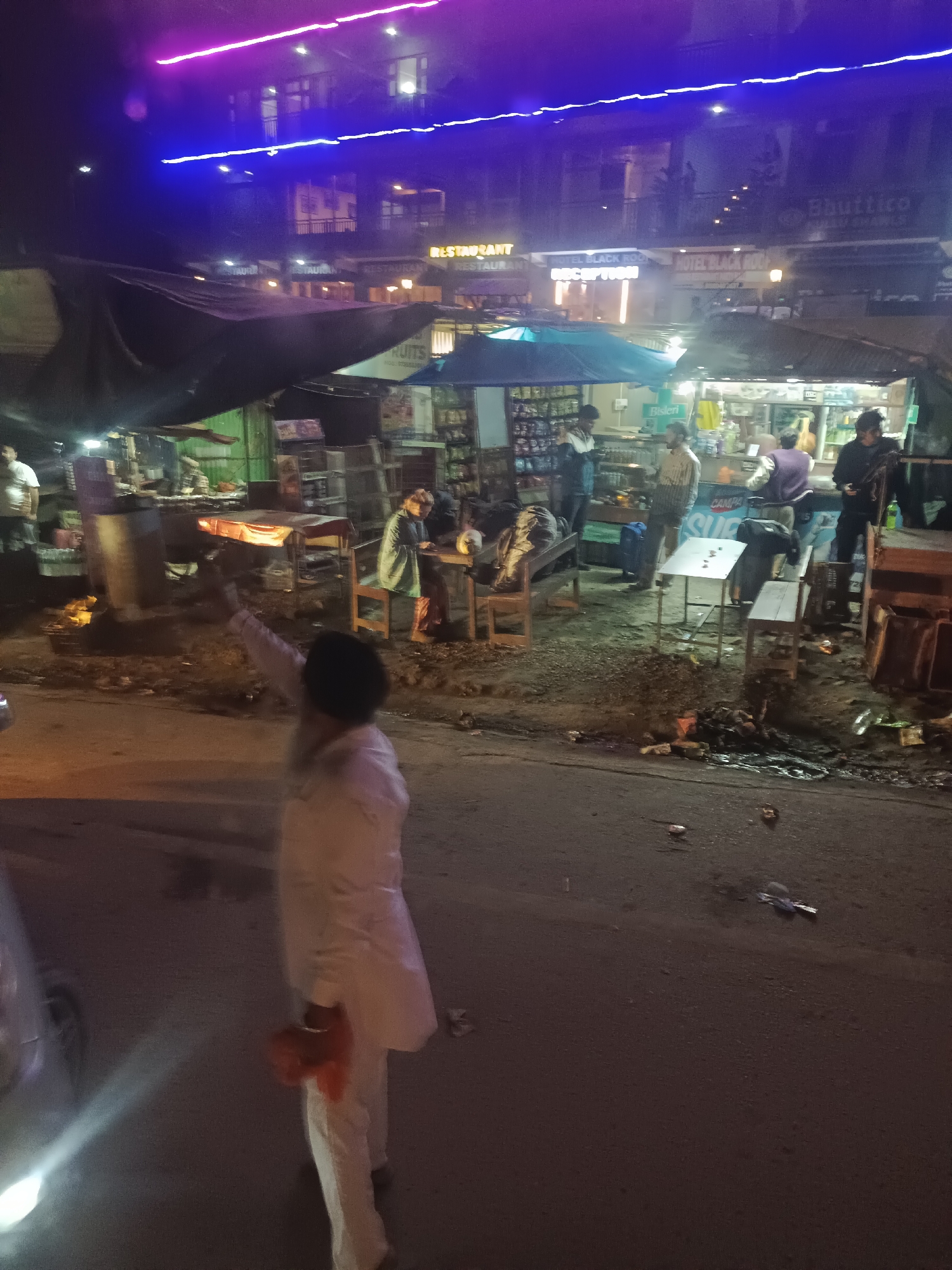 A nighttime street scene in Bhuntar, India, is depicted with a focus on a small marketplace area. The foreground shows a person in white clothing holding a red object. The middle ground features several makeshift stalls with goods displayed, some under tarps. A few individuals are visible interacting within these stalls, including one person behind a counter. Wooden tables and chairs are set up in front of the stalls. The background reveals a multi-story building with illuminated windows and signage that includes "HOTEL BLACK ROCK." Neon lights, one purple and one blue, are strung across the building's facade. The street itself appears dirty, with scattered debris. The overall atmosphere is that of a busy, dimly lit commercial area at night.