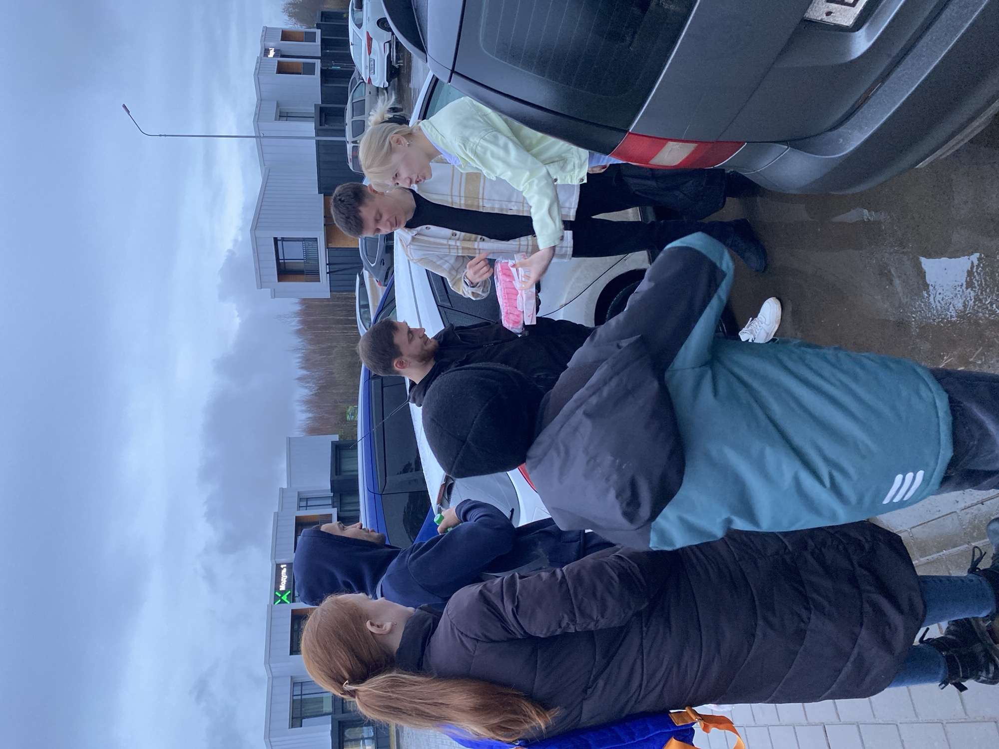 A group of six people stand near a row of parked cars in Votkinsk, Russia.
Several individuals are gathered around the open trunk of a white car, with a woman in a yellow jacket and a man in a plaid shirt looking at a pink package.
Another group of people stands nearby, with one individual wearing a blue hooded jacket, and another in a black jacket with red hair in a ponytail. A person with a black beanie is also present.
The cars are parked in front of a building that has a sign in Cyrillic. The sky is overcast, and the ground appears wet.