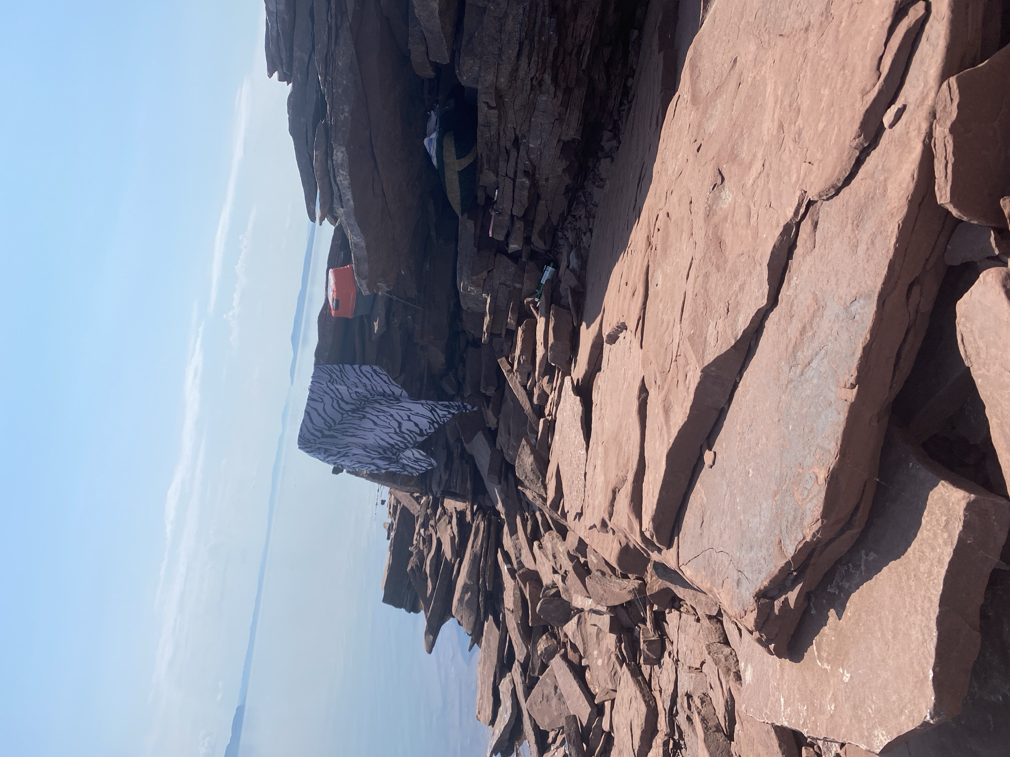 A low-angle, full shot captures a rocky shoreline with a serene, expansive body of water under a clear, pale blue sky. The foreground is dominated by weathered, reddish-brown rocks, some flat and layered, others broken and scattered, creating a rugged texture. In the middle ground, a series of layered rock formations jut out towards the water. A white fabric with a black zebra-like pattern is draped over one of these ledges. To the right of the fabric, an orange cooler rests on the rocks. Further back and slightly to the right, nestled within a rocky overhang, is a green and yellow striped sleeping bag. A few scattered bottles, one appearing to be green and another clear with a white label, are visible amongst the rocks in the overhang. The water is calm and reflects the pale sky, with distant landmasses or mountains visible on the horizon. The scene appears to be in a natural, outdoor setting, possibly a secluded spot by a lake or sea. No people are present in the image. The location is identified as Ust'-Erba, Russia.