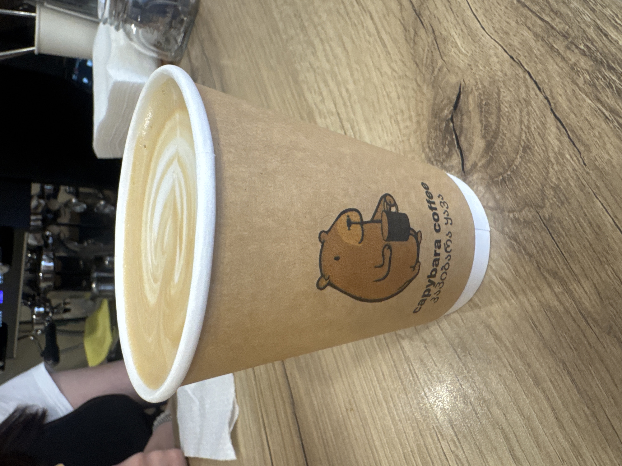 A latte in a paper cup with a capybara logo sits on a wooden table. The latte has a white foam design. The background shows a coffee machine and someone wearing a white shirt. The cup has "capybara coffee" and Georgian text printed on it. The scene appears to be inside a coffee shop in the city of Unknown, Unknown.
