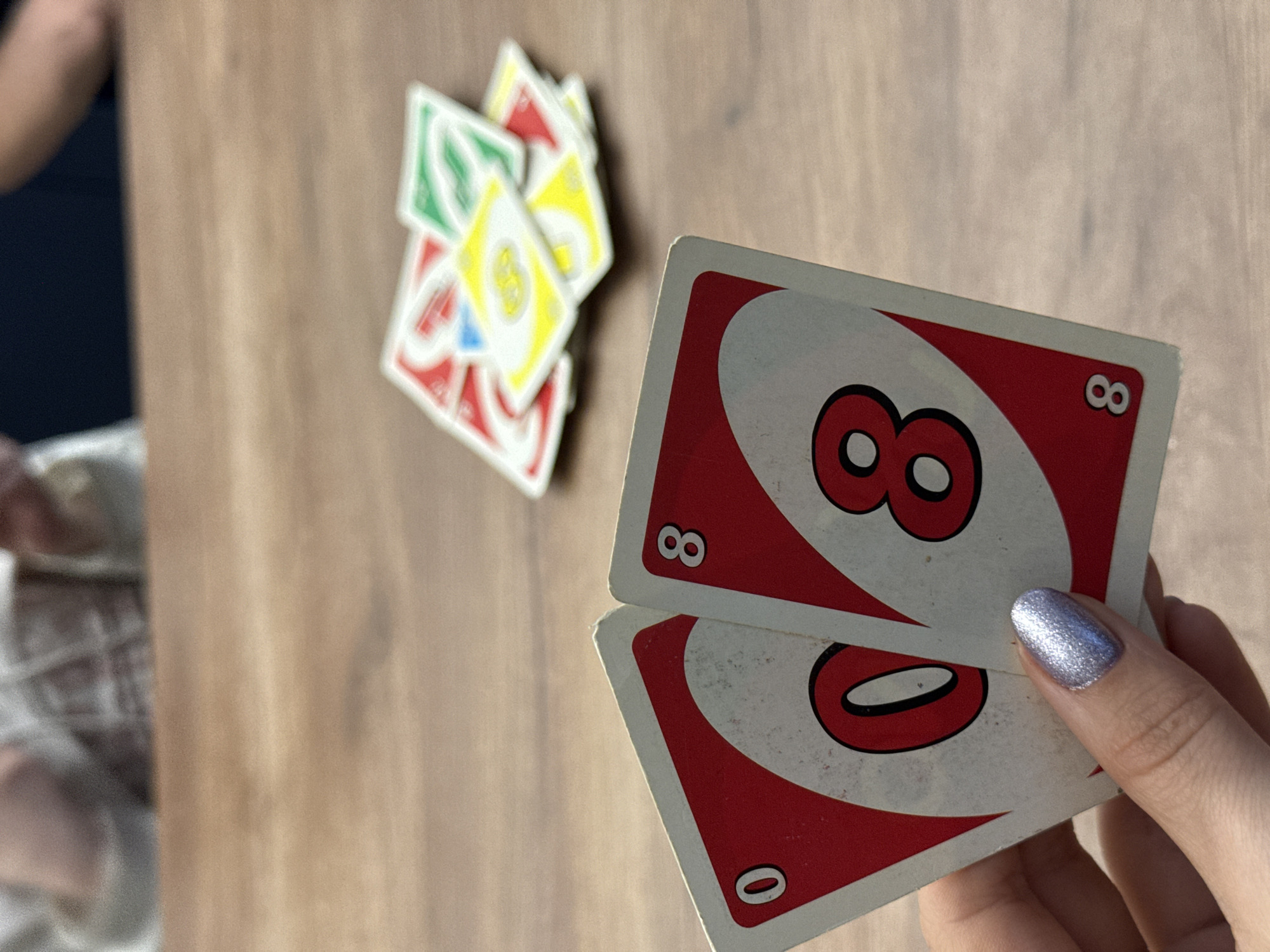 A close-up photograph shows a person holding two Uno cards: a red '8' and a red '0'. Several other Uno cards are blurred and scattered on a wooden surface in the background. The photograph does not show any identifiable features to indicate that it was taken in Лобаново, Russia.