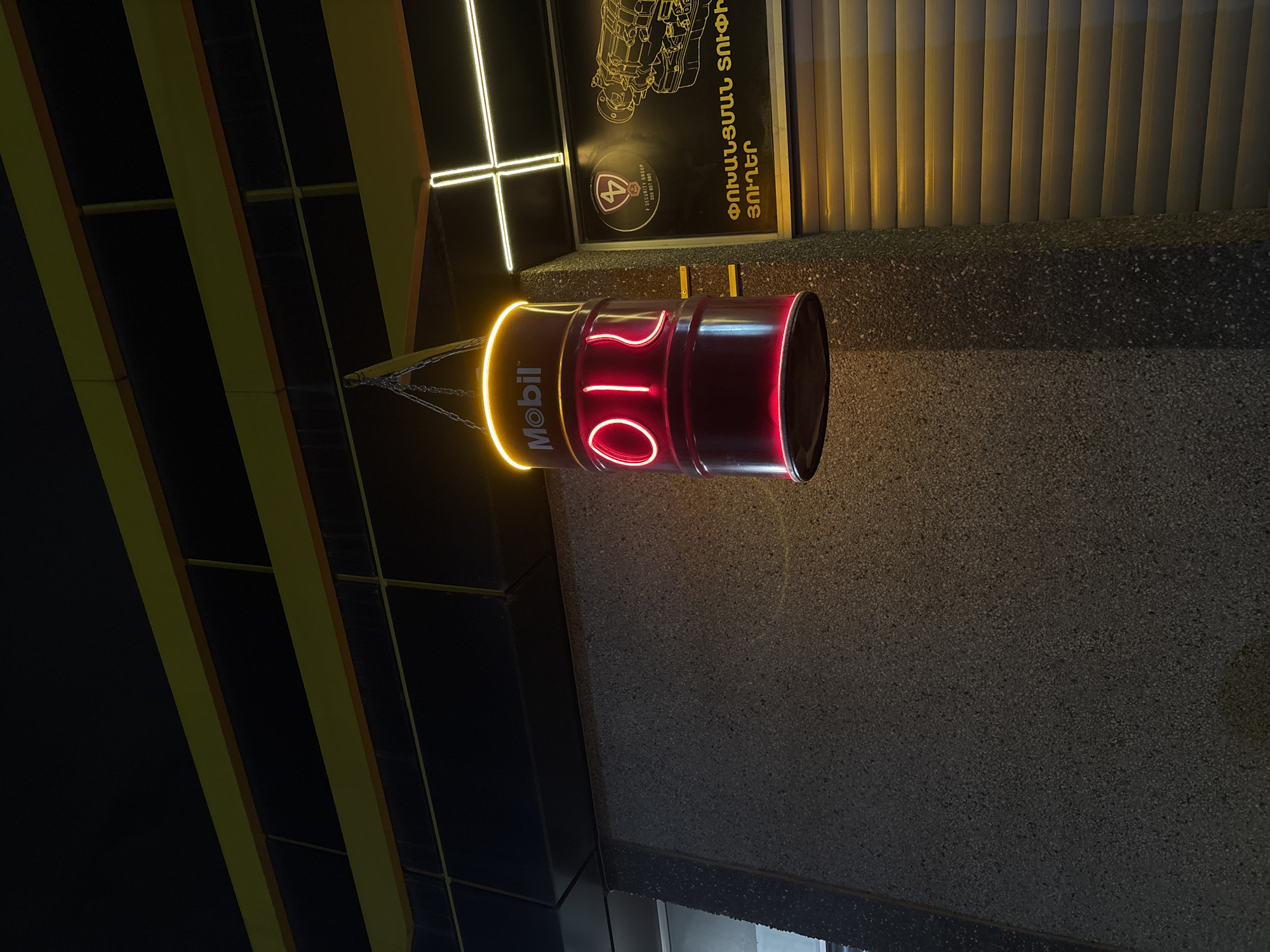 Here is a description of the image:
The image shows a Mobil oil barrel sign suspended by chains from a dark ceiling. The barrel is cylindrical, with the word "Mobil" illuminated in yellow on one side. On the other side, the numbers "010" are displayed in red neon lighting. The background includes a textured wall and a sign with Armenian text and a logo. A neon cross is affixed to a sign. This sign is in front of window blinds.
The picture was likely taken in the city of Yerevan, Armenia, as indicated by the Armenian text on the nearby sign. The presence of an oil barrel and a shop sign suggests the location may be in front of or near an auto shop.