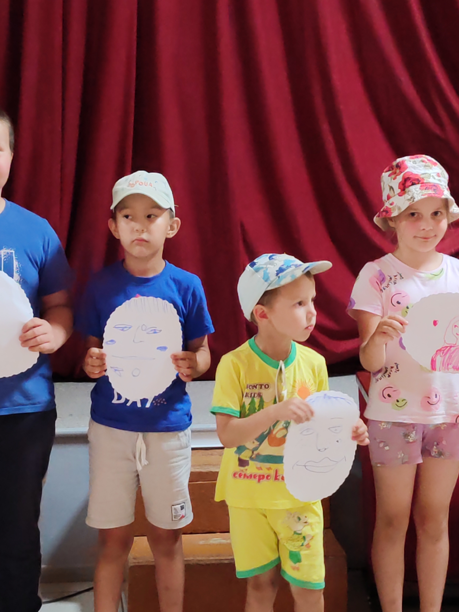 In the city of Chukhlom, Russia, four children are posing for a photograph in front of a red curtain. On the left, a child wearing a blue t-shirt and black pants holds a white circular object. Next to them, a boy in a blue t-shirt and light-colored shorts holds a white paper cutout of a face with drawn features. To his right, another boy in a yellow t-shirt and yellow shorts with green trim holds a similar white paper cutout of a face. On the far right, a girl in a floral-patterned hat and a pink t-shirt with smiley faces, wearing pink patterned shorts, holds a white circular object with a drawing on it. The children appear to be participating in an art or performance activity.