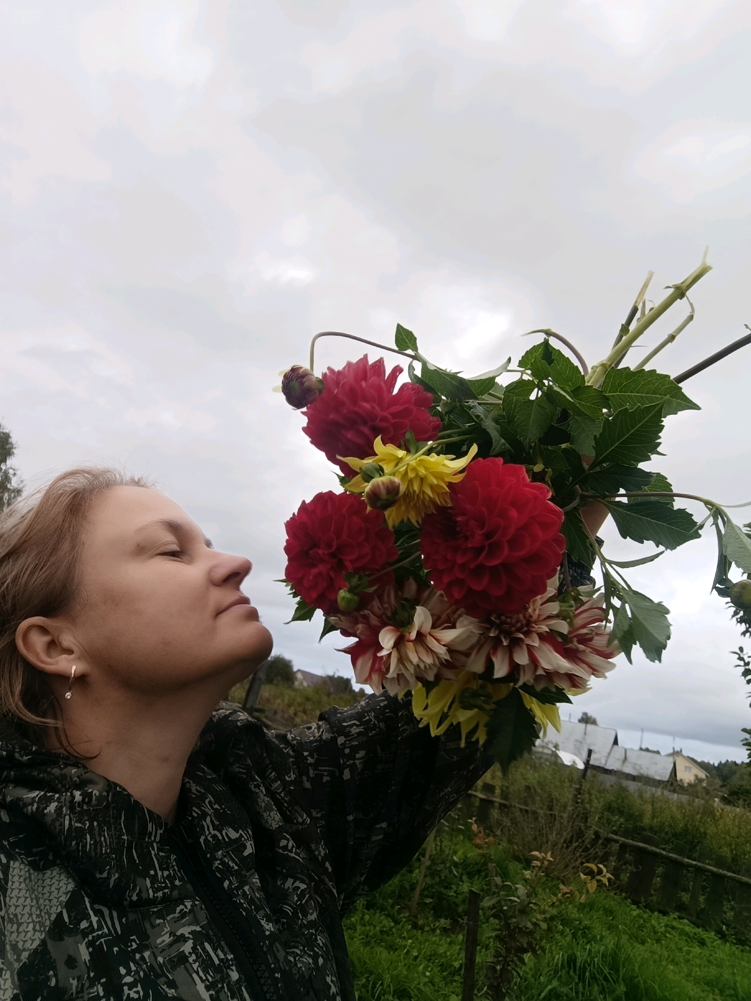 A woman with blonde hair is holding a bouquet of red and white dahlias and yellow flowers up to her face, as if to smell them. She is wearing a dark camouflage jacket. The background shows a rural landscape with green grass, trees, a wooden fence, and houses with gray roofs. The sky is cloudy and overcast. The scene appears to be in Dubovaya, Russia.